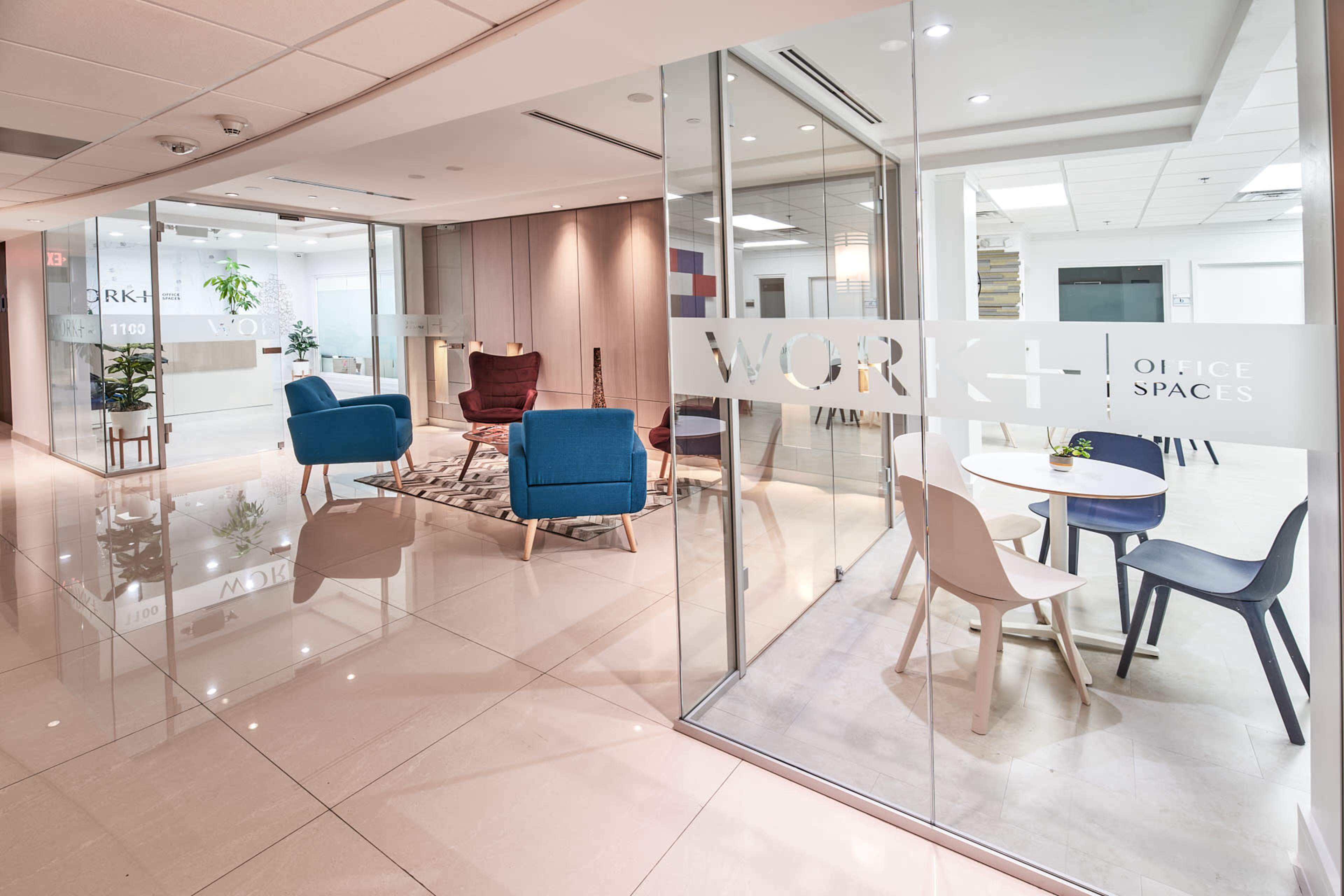 A modern office space featuring glass partitions, a seating area with blue and burgundy chairs, and a small round table in a bright, well-lit environment.
