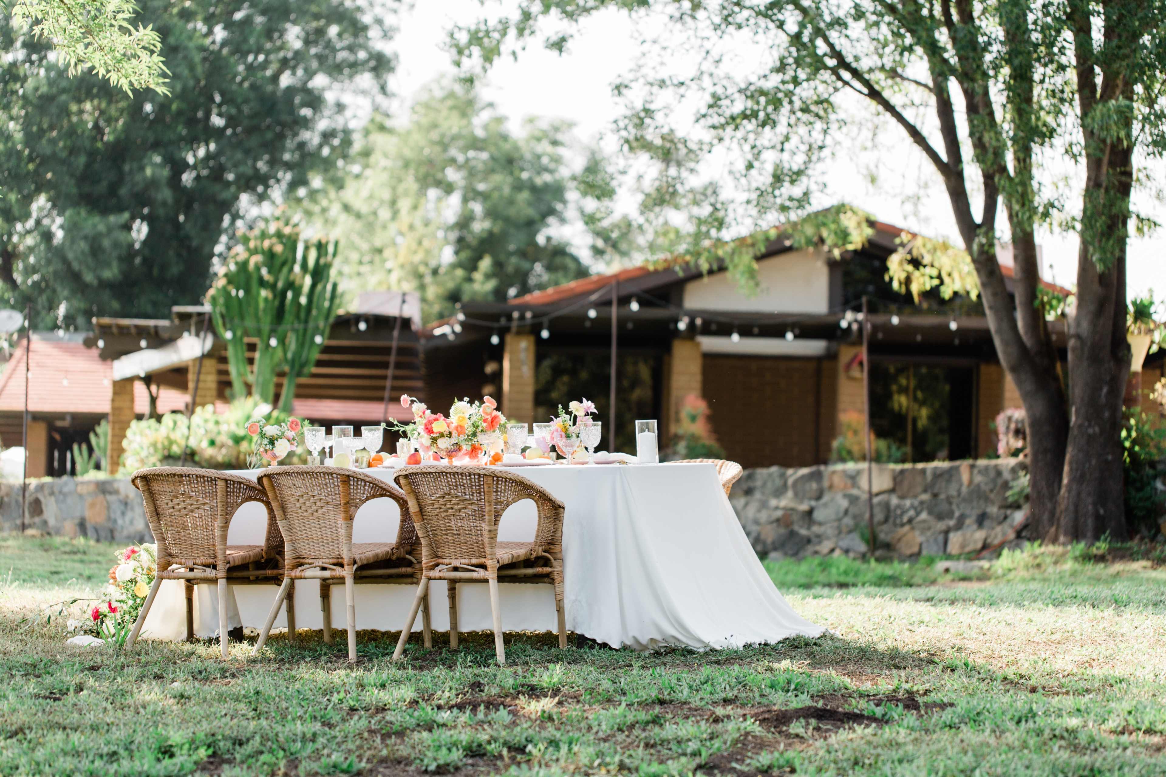 A beautifully set outdoor dining table with wicker chairs is positioned on grass in front of a house surrounded by trees.