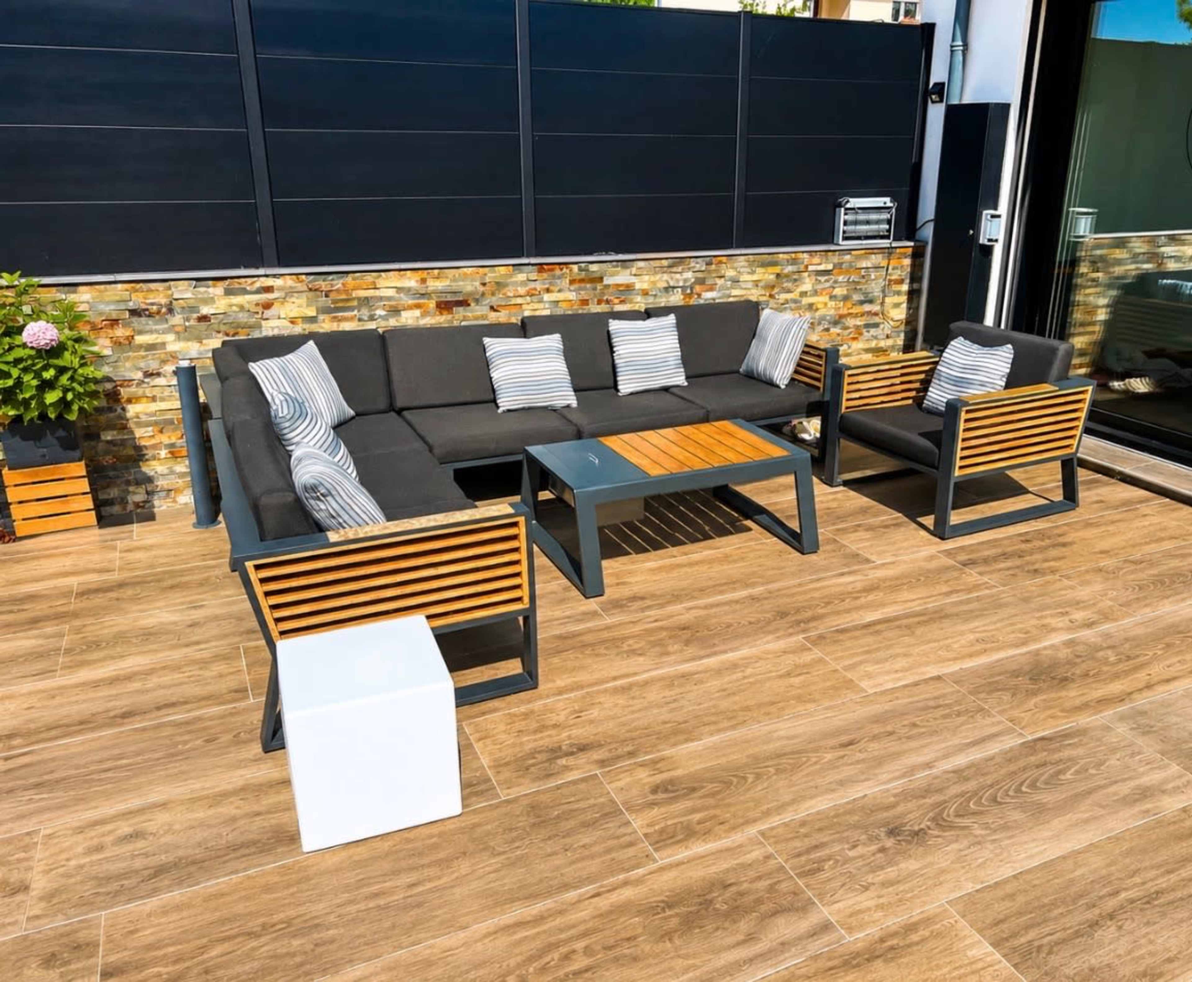 The image shows a modern outdoor living space with a large sectional sofa, a coffee table, and additional seating arranged on a tiled patio.