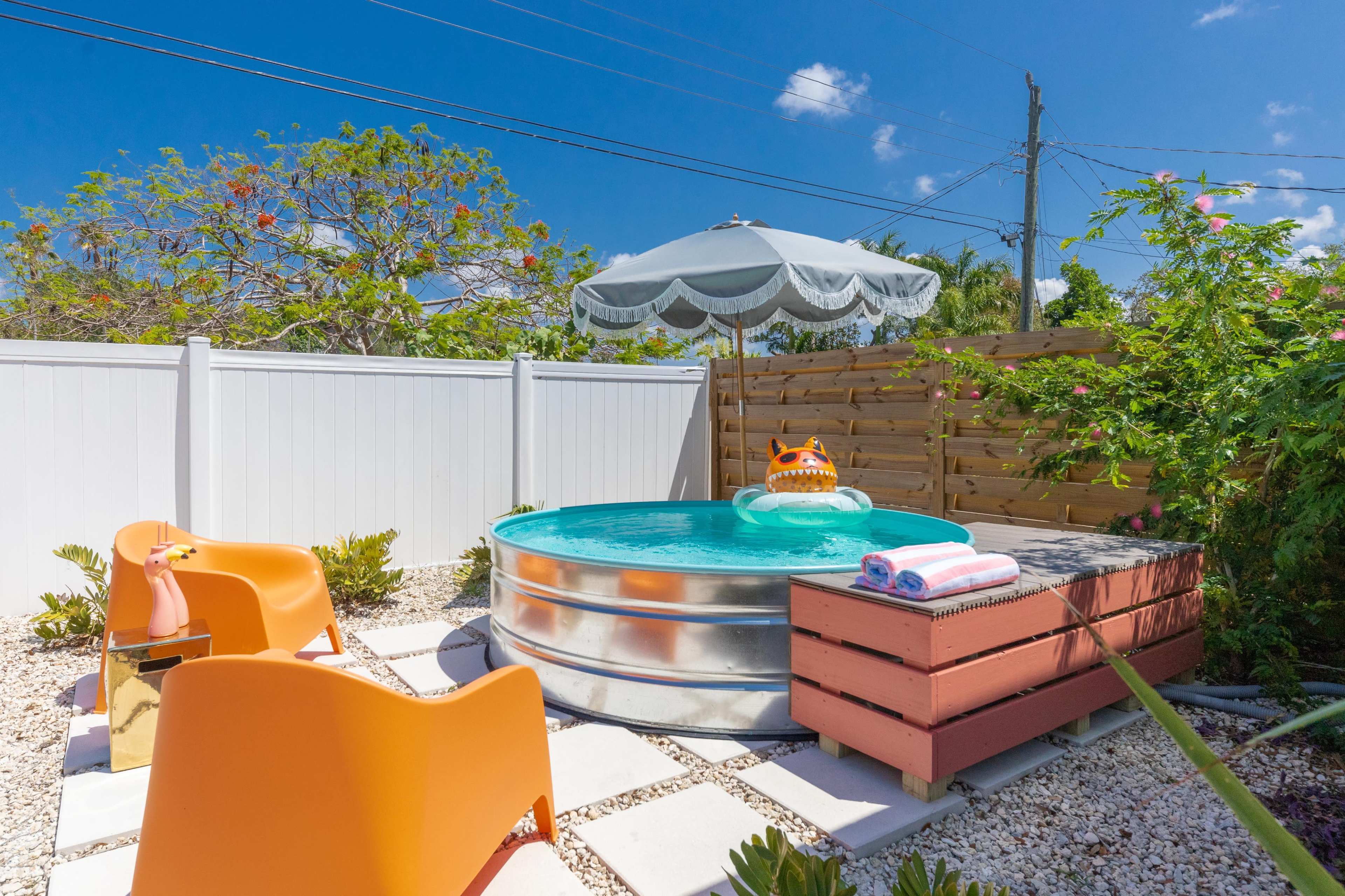A small metal pool surrounded by stone pavers, two orange chairs, a wooden box, and a shaded umbrella in a fenced outdoor space.