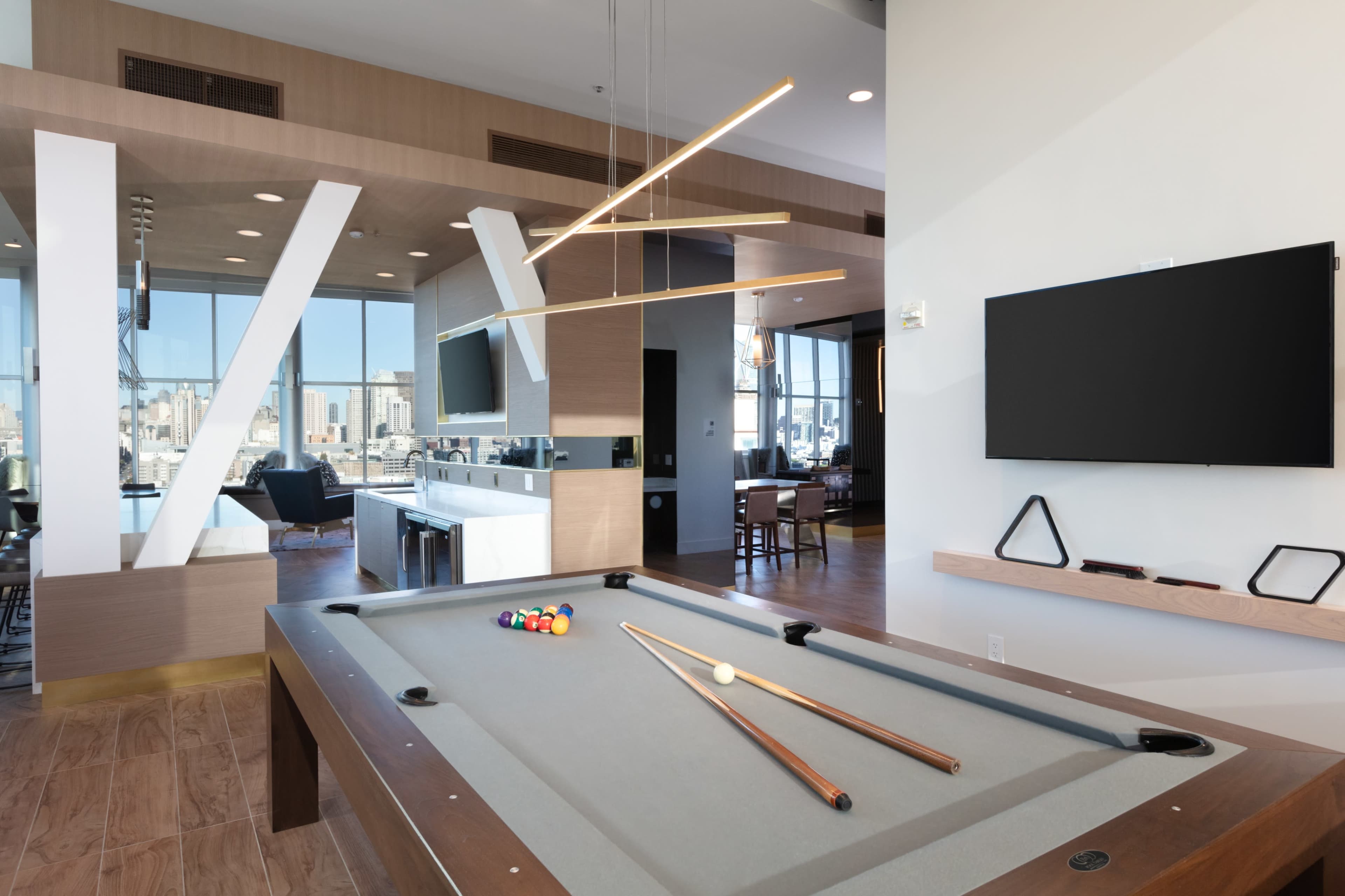 A modern game room featuring a pool table, a sleek entertainment area with a large TV, and large windows that provide a view of the city.