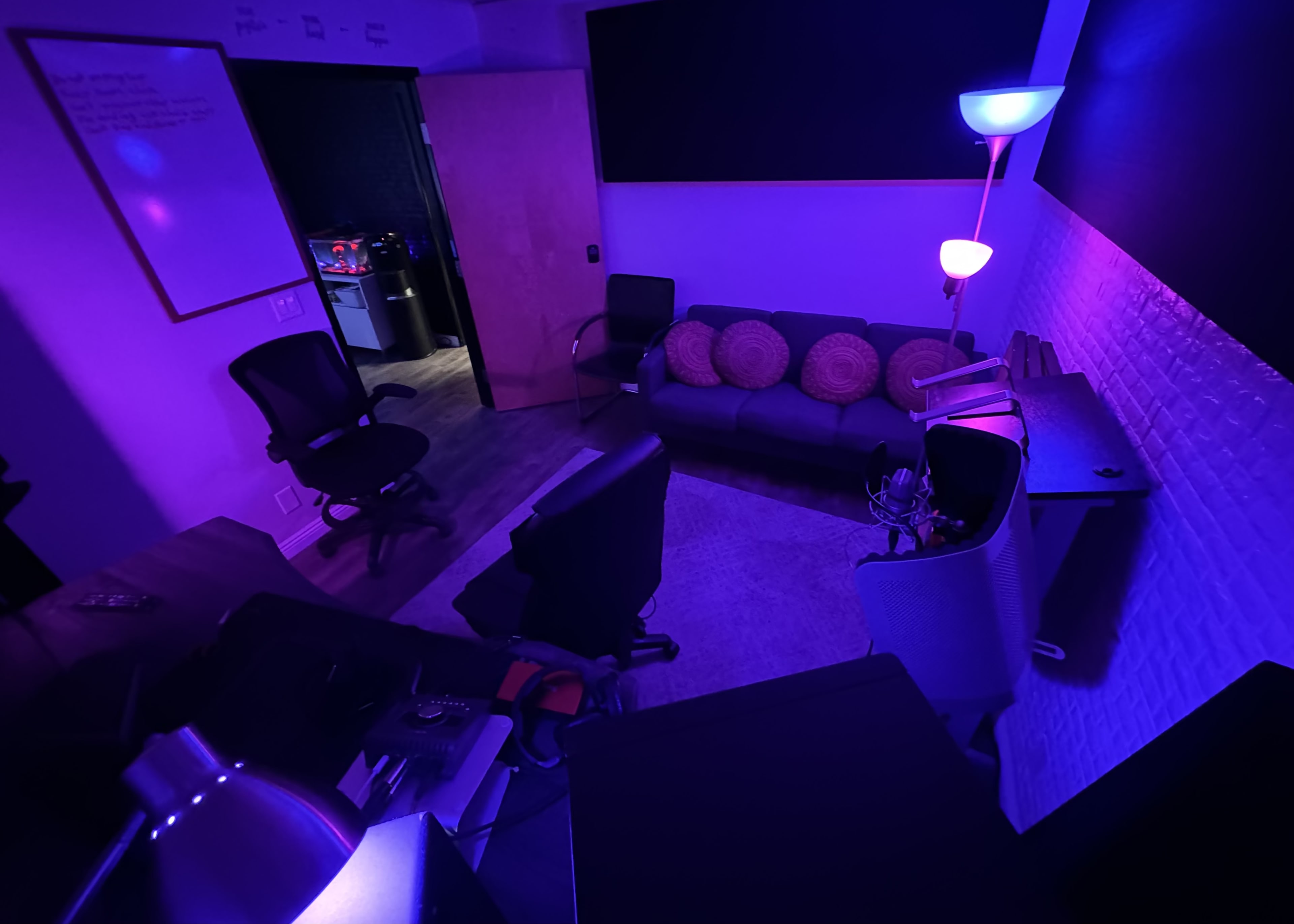 A dimly lit room featuring a couch, office chairs, and a standing lamp with purple light.