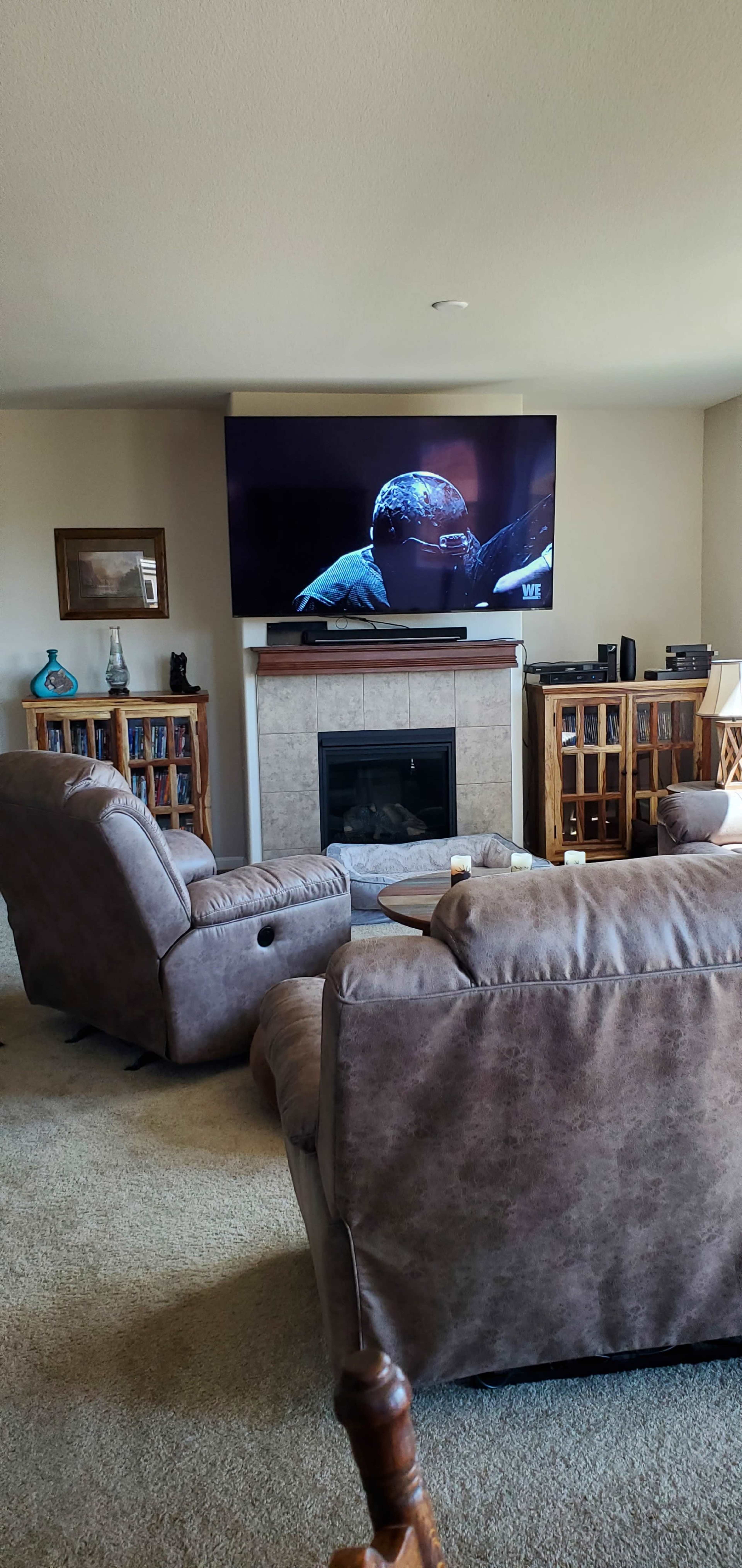 A living room featuring two recliner chairs facing a mounted television above a fireplace, with bookshelves and audio equipment in the background.