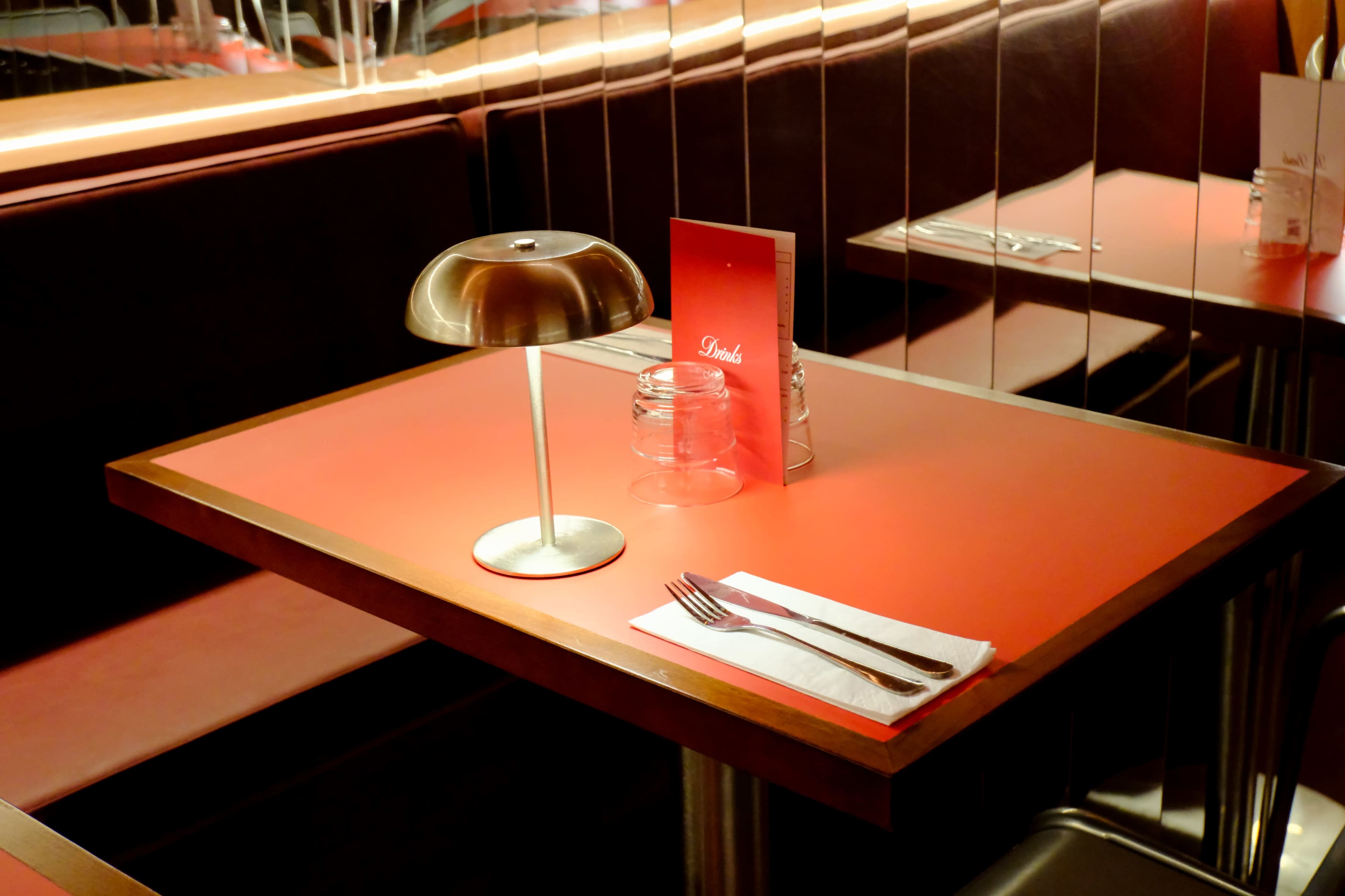 A red-topped table with a metal lamp, a glass jar, and a menu sits in a restaurant booth.