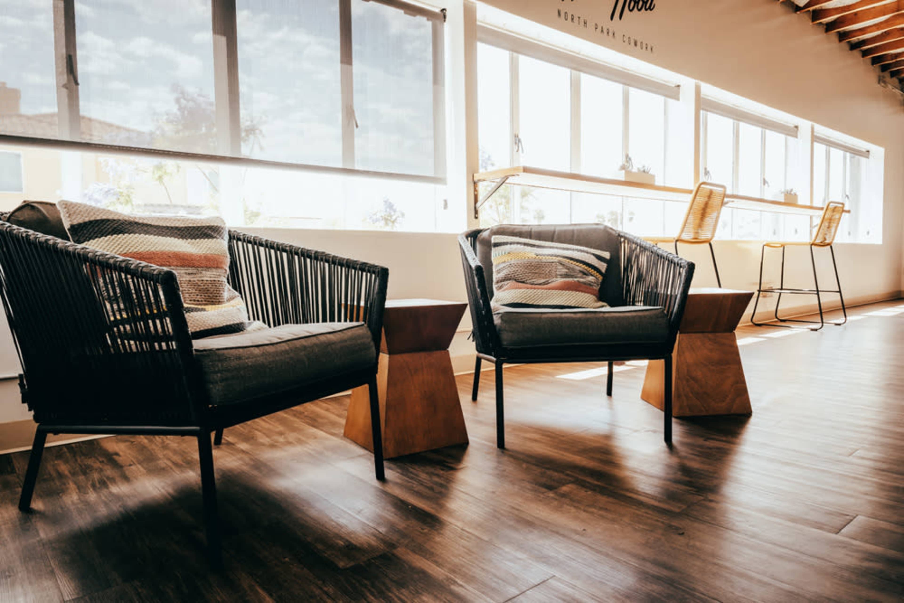 Two woven armchairs with patterned cushions sit beside wooden side tables in a bright room with large windows.