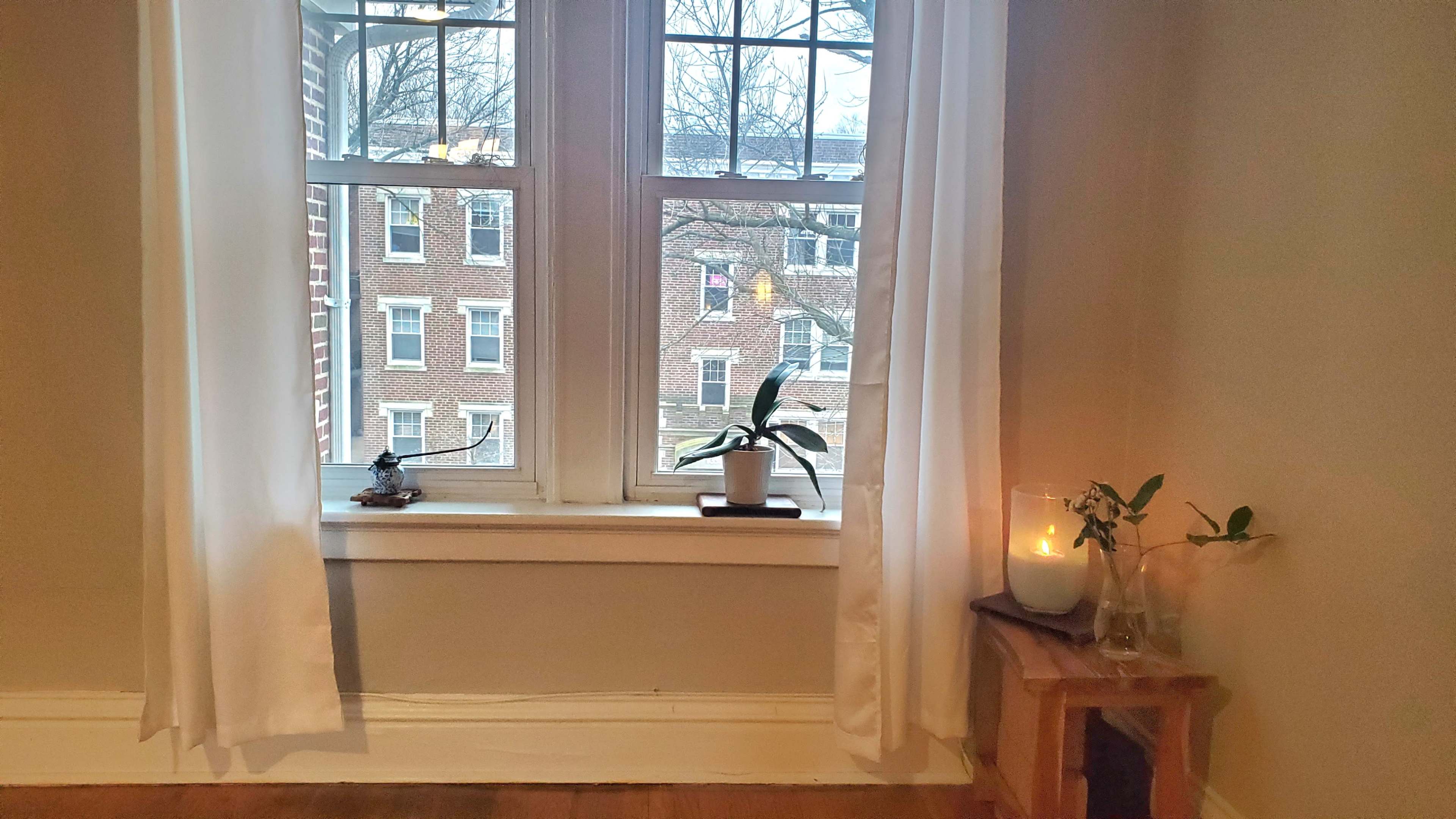 The image shows a light-filled room with two large windows, a potted plant on the windowsill, a candle, and a small wooden table beside a vase with flowers.