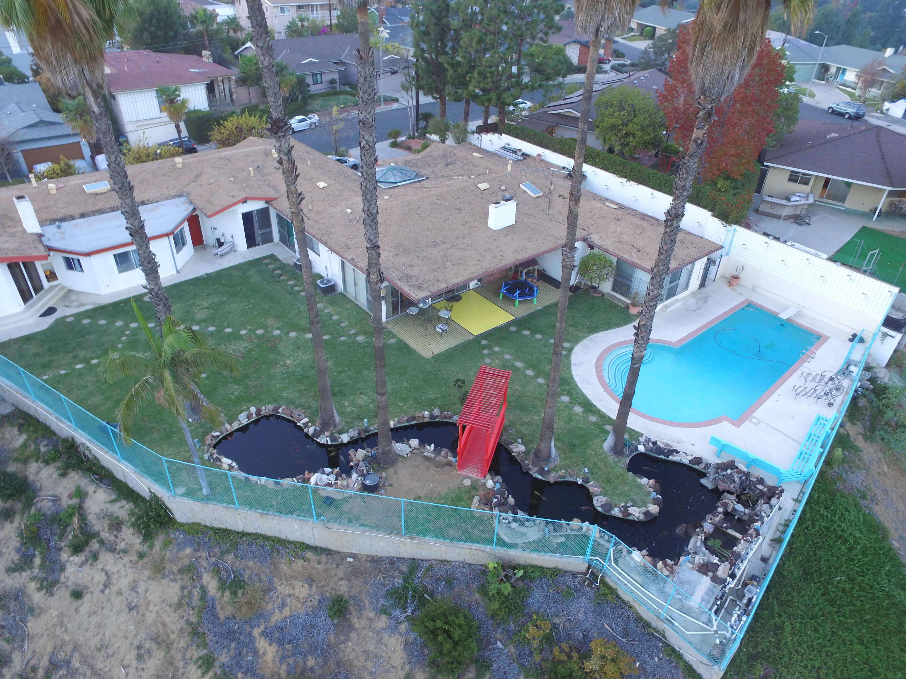 The image shows a single-story house surrounded by palm trees, featuring a swimming pool and a landscaped backyard with a small pond and a decorative red structure.