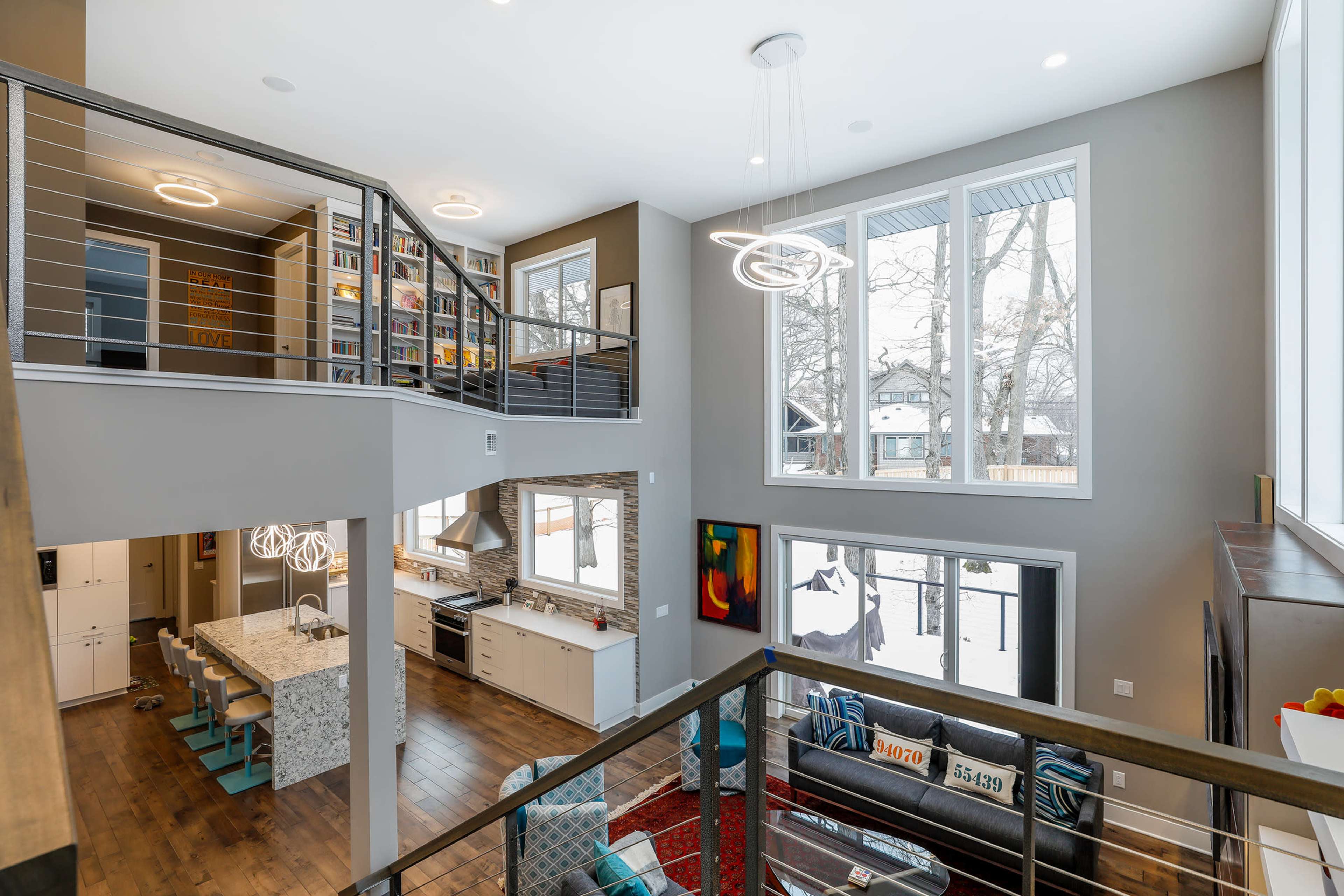 A modern two-story living space with large windows, a sleek kitchen, and a balcony overlooking a lower level furnished with a colorful rug and art.