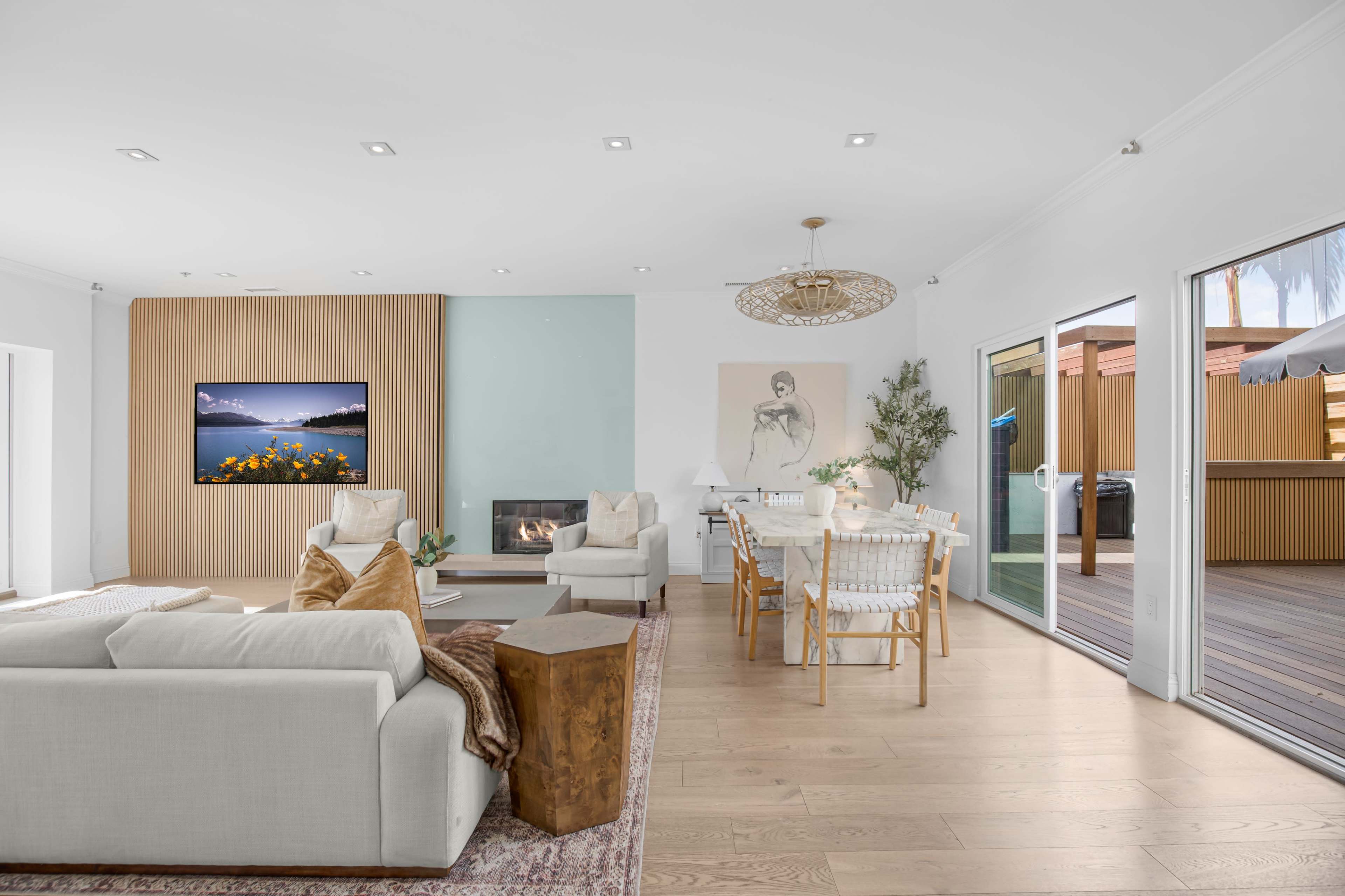 The image shows a modern living space featuring a light-colored sofa, a wooden accent wall with a television, a dining area with a table and chairs, and large glass doors leading to an outdoor deck.