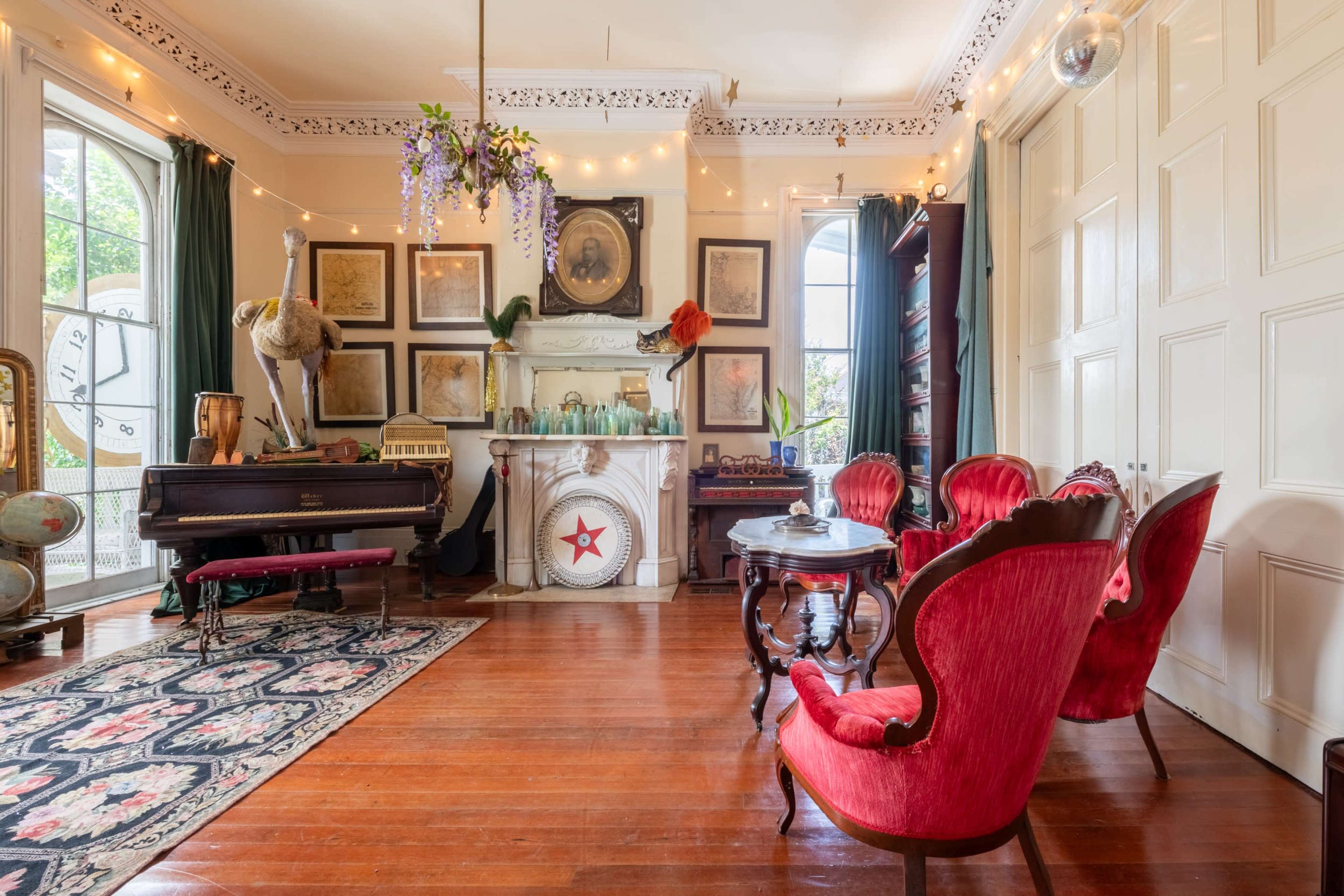 The room features a vintage piano, ornate red armchairs, and a fireplace adorned with framed artwork and decorative items, all under soft lighting from string lights.