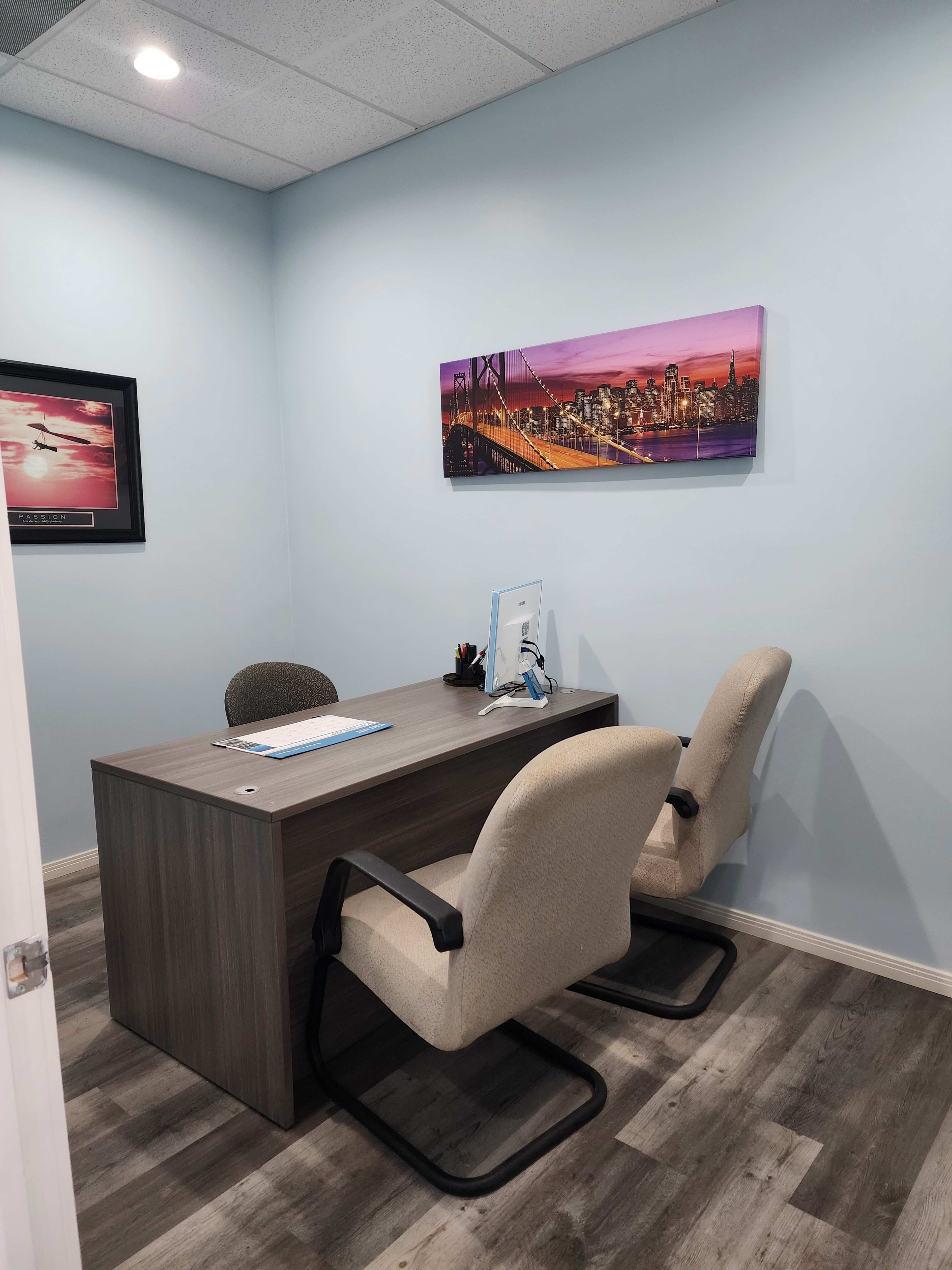 The image shows a small office space with a wooden desk, two chairs, and a colorful cityscape photograph on the wall.
