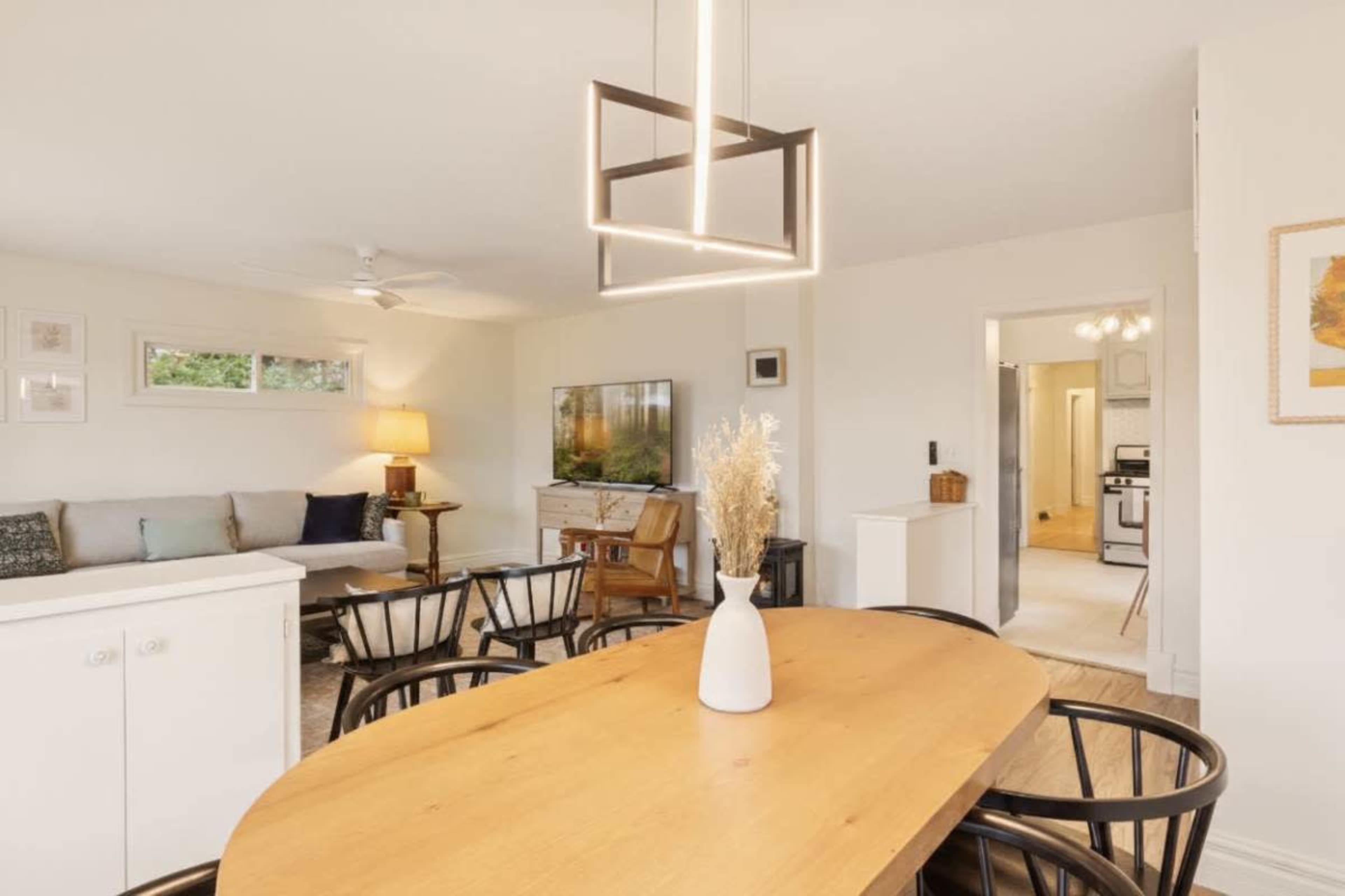 The image shows a bright living space with a dining table at the center, surrounded by black chairs, and a seating area with a sofa and TV in the background.