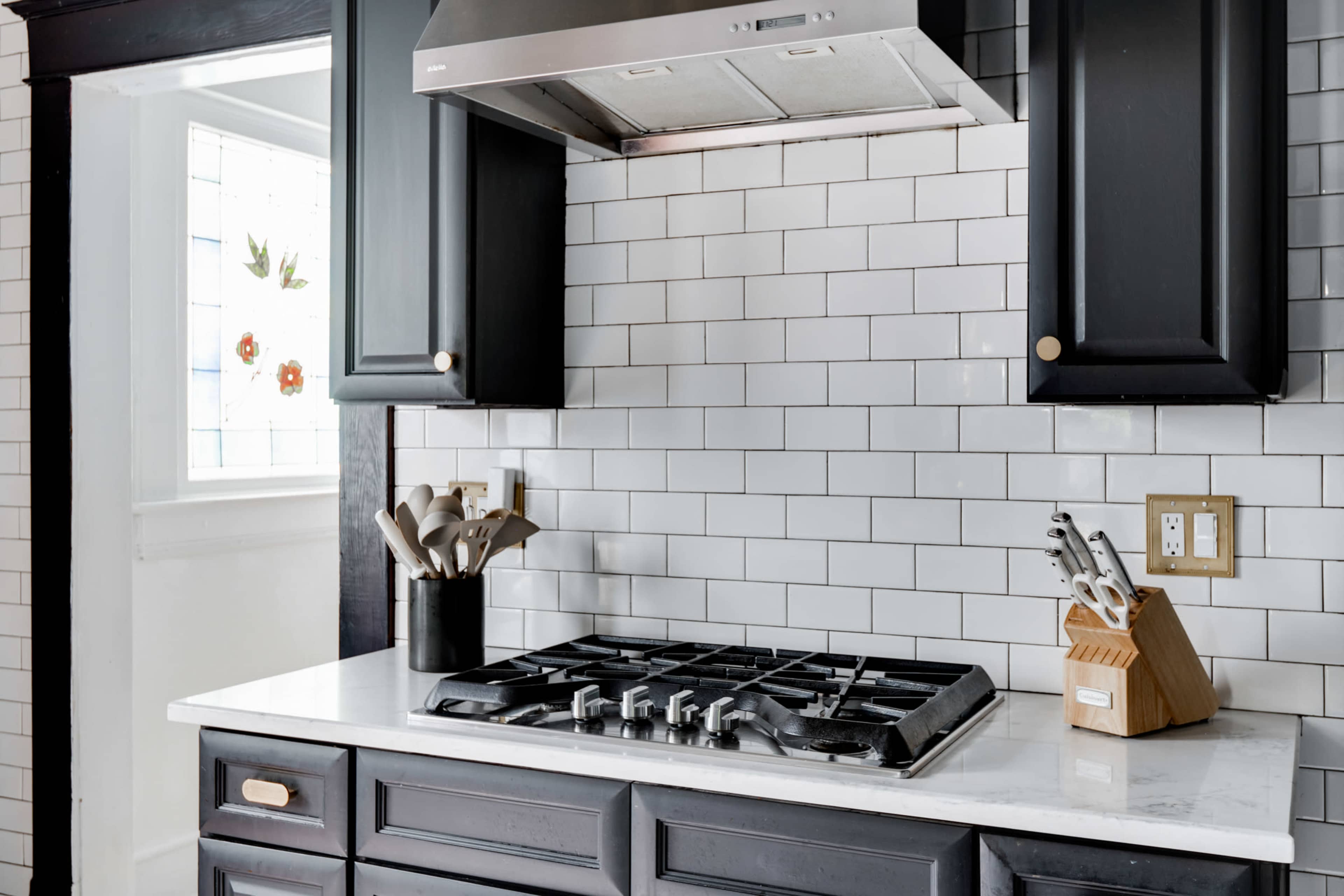 The image shows a modern kitchen with a gas stove, a stainless steel range hood, white subway tile backsplash, and dark cabinetry.