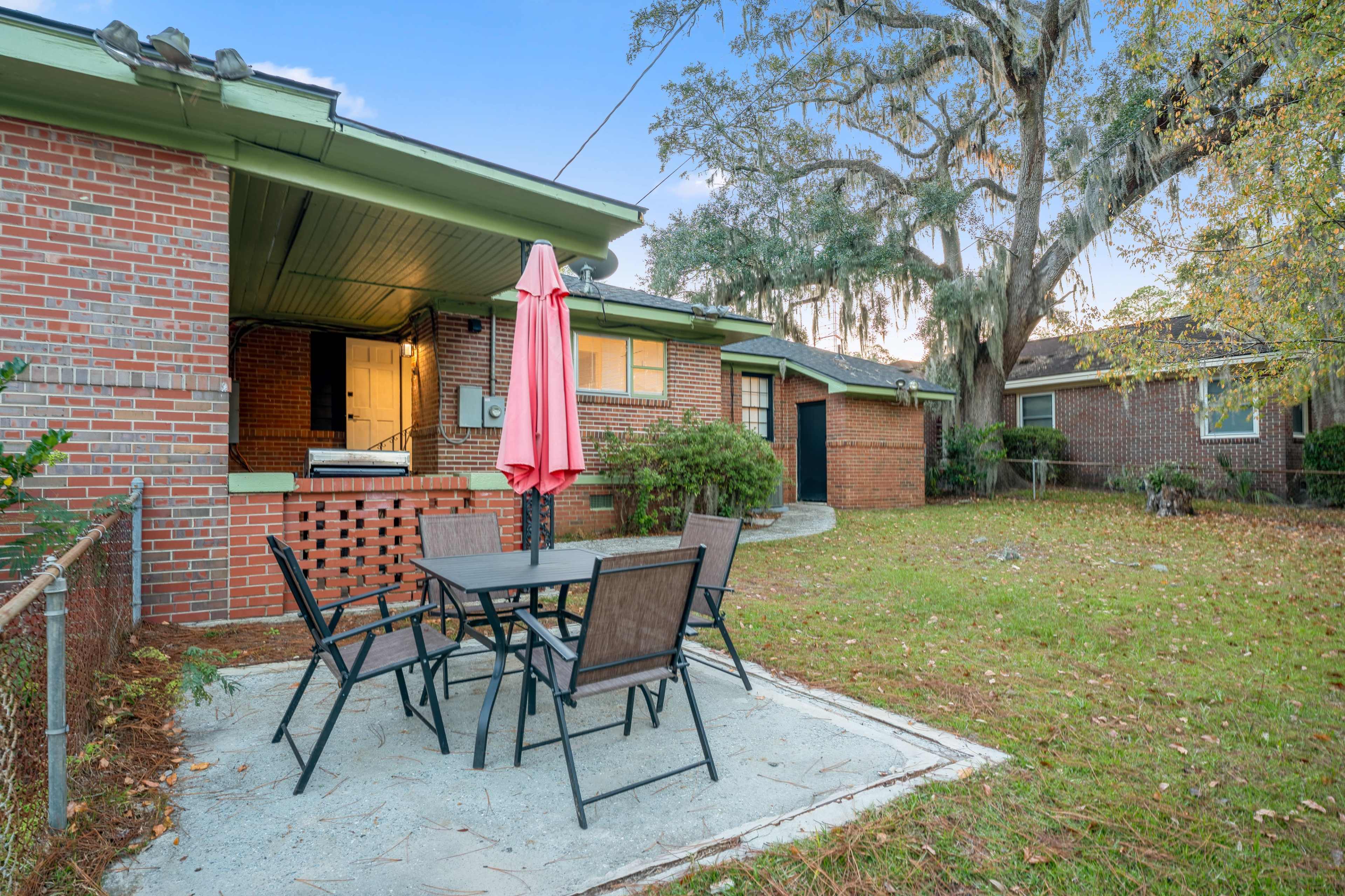 The image shows an outdoor patio area with a table and chairs covered by a pink umbrella, surrounded by grass and trees near a brick home.