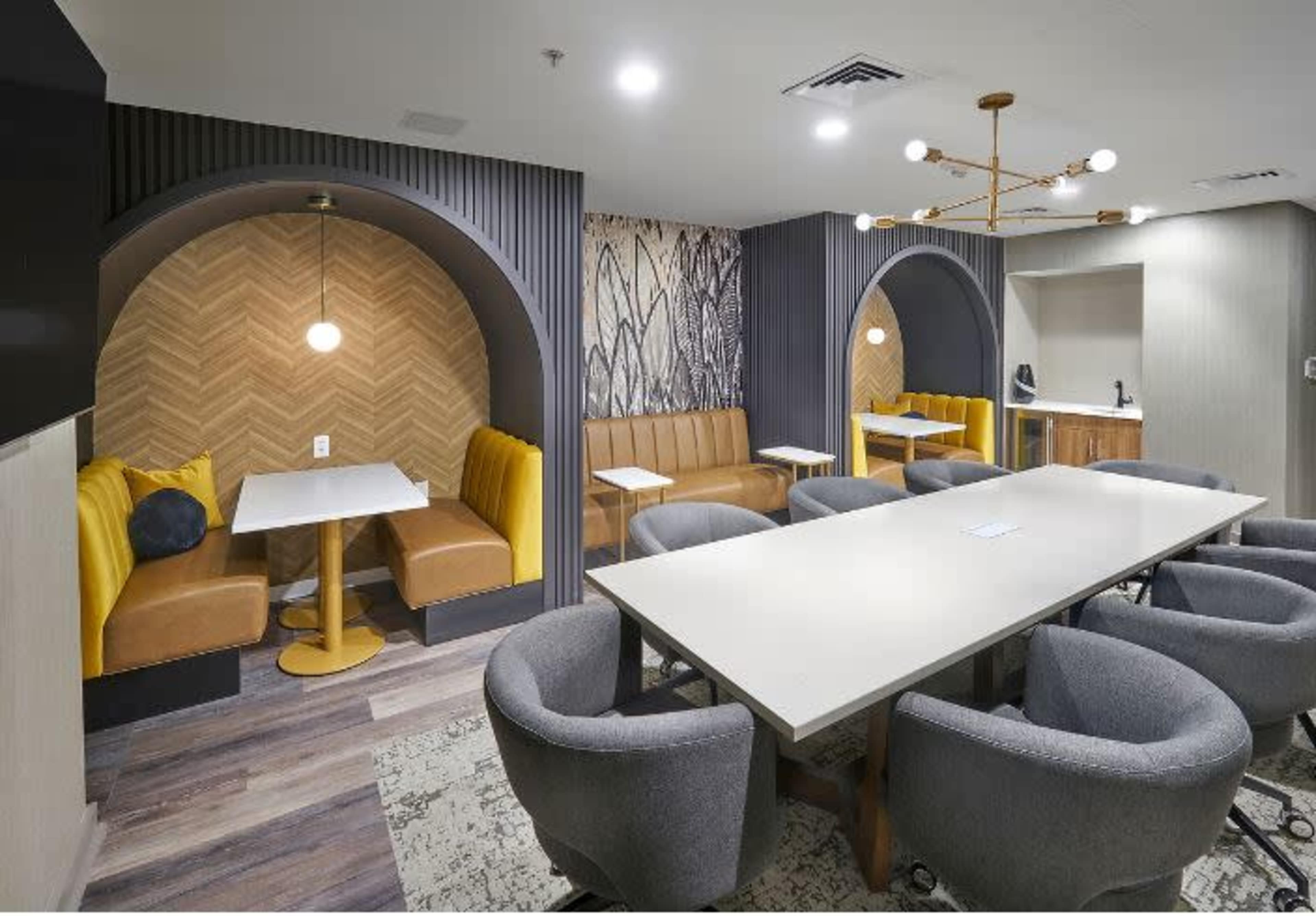 The image shows a modern meeting area featuring two semi-private seating alcoves with yellow upholstered benches and a central table surrounded by gray chairs.