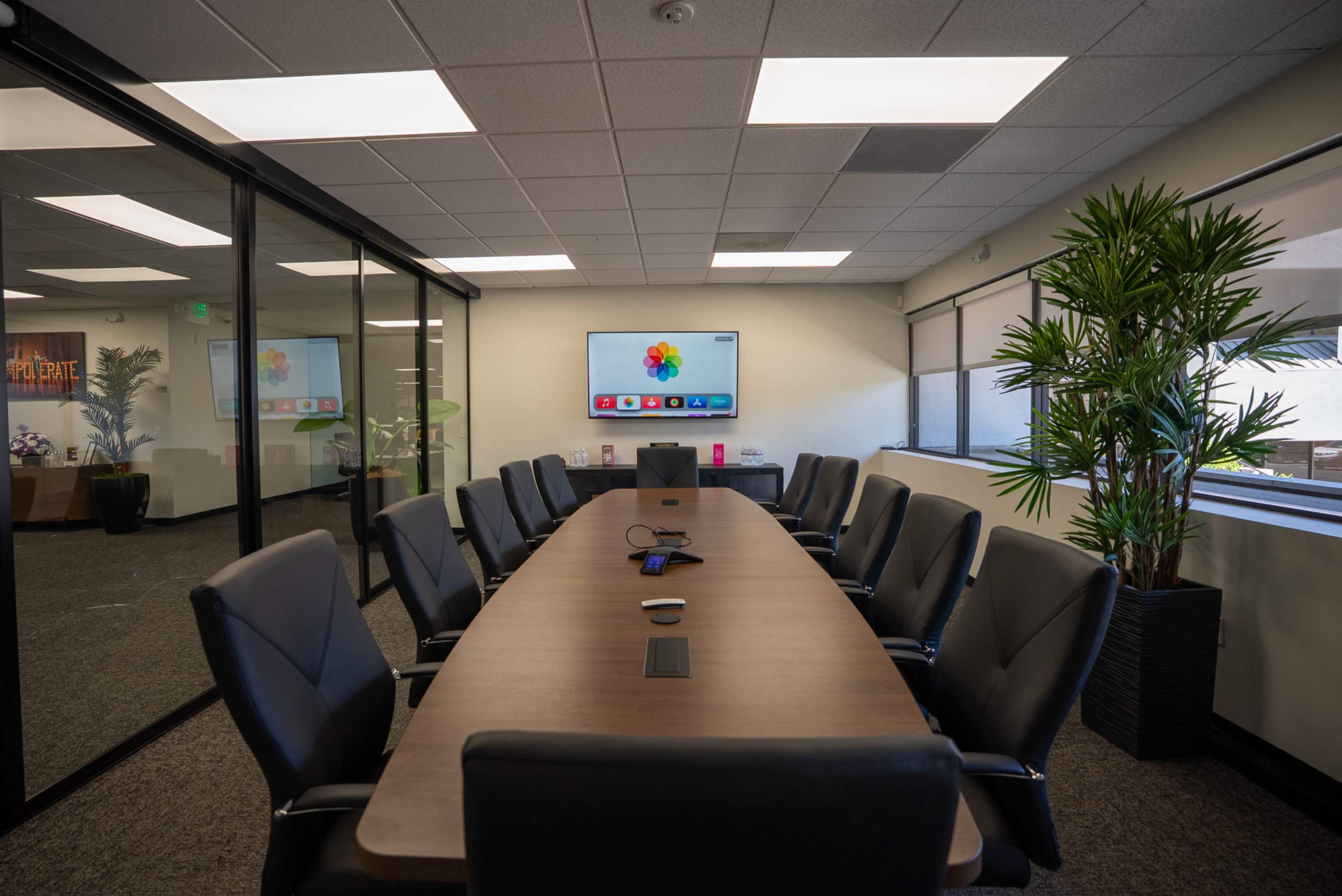 A large conference room features a long wooden table surrounded by black chairs, with a wall-mounted screen displaying colorful icons.