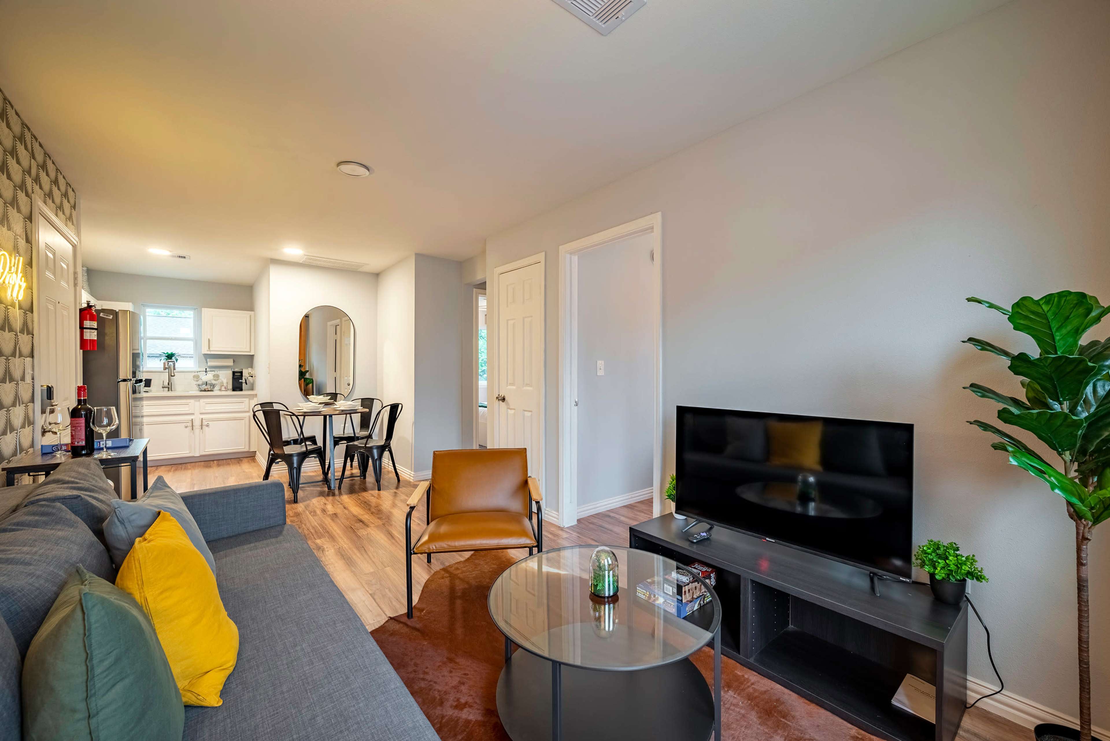 A modern living area features a gray sofa, a glass coffee table, and a television opposite a kitchenette with dining space.