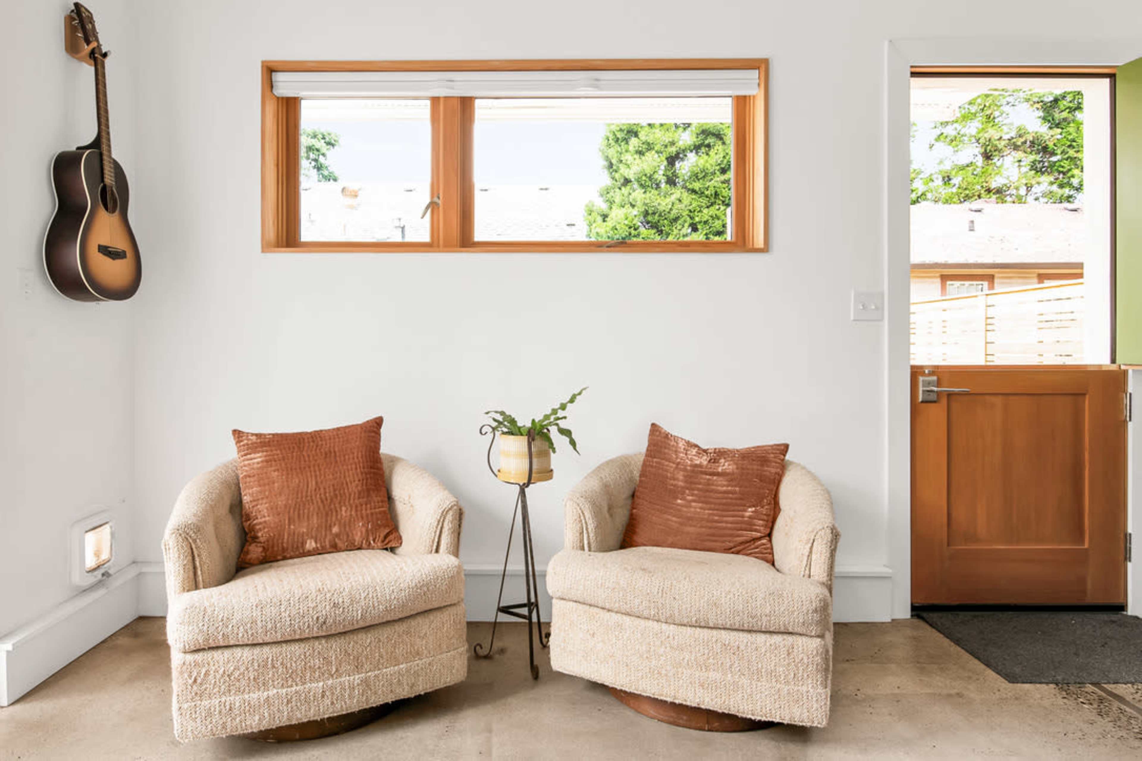A bright interior space with two cozy chairs, a small side table with a plant, and a guitar hanging on the wall.