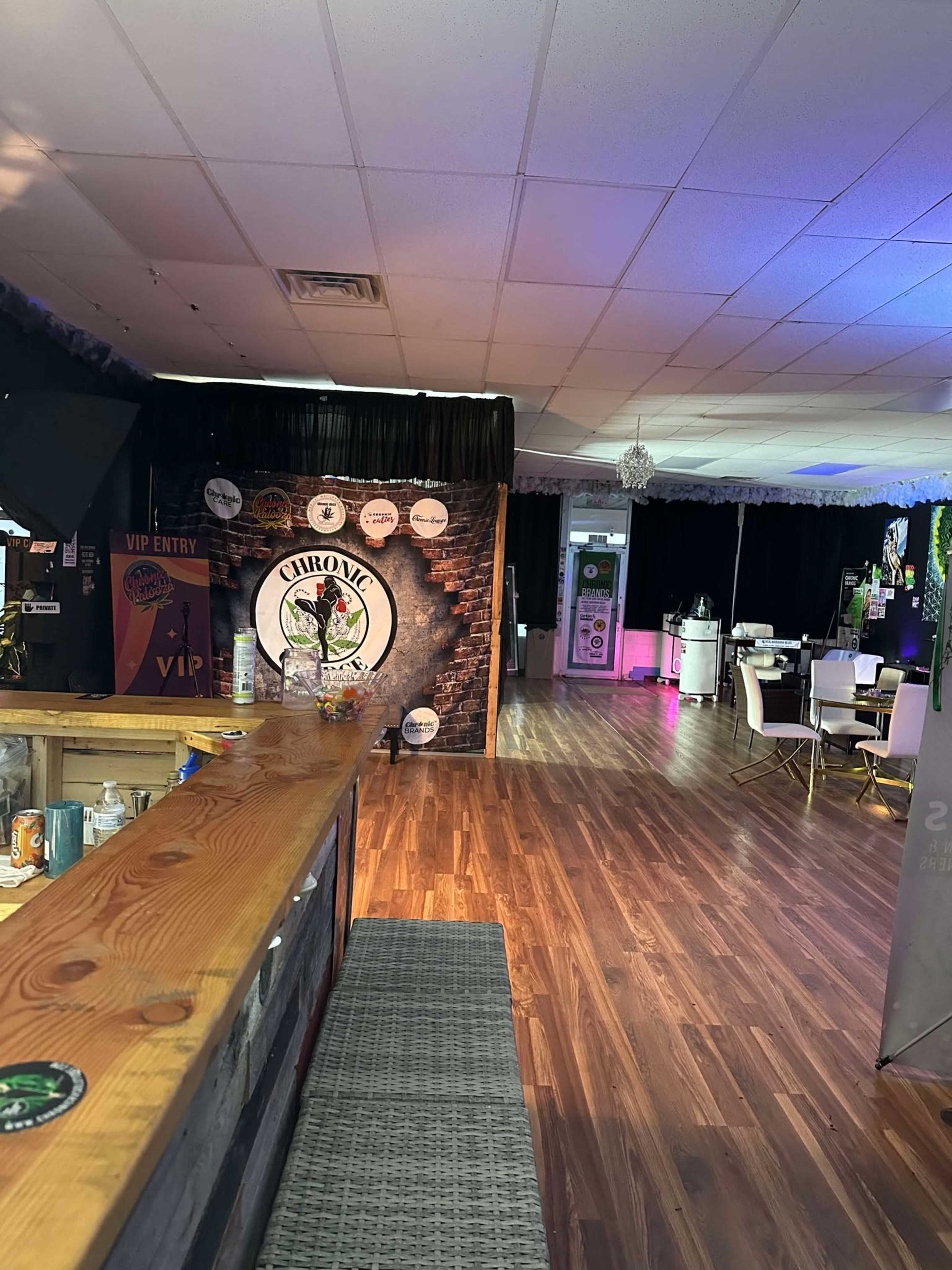 The image shows the interior of a venue with a wooden bar, a promotional backdrop featuring the word "Chronic," and seating areas lit with colored lights.