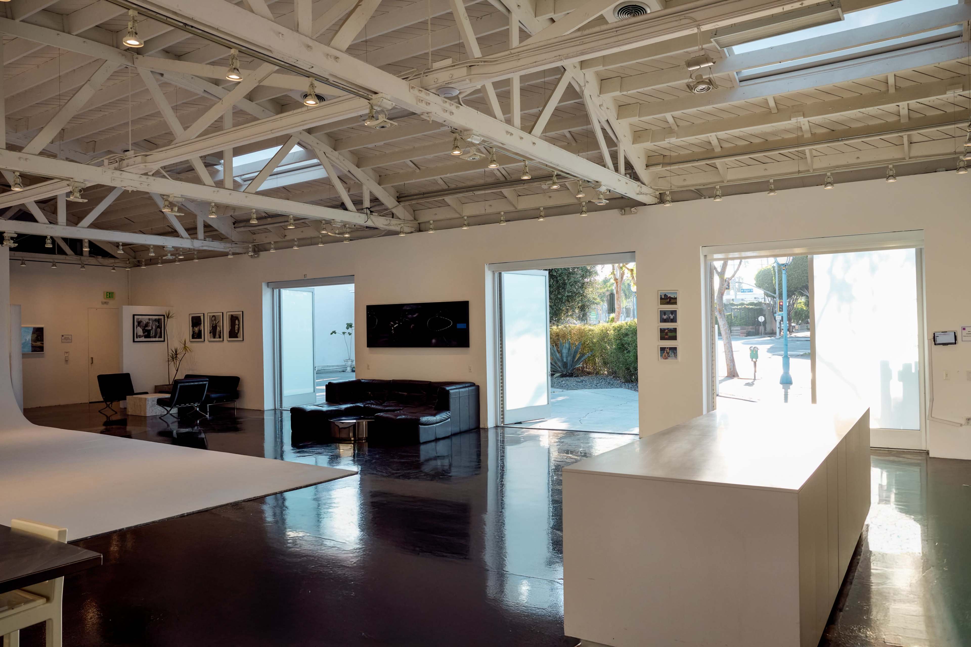 The image shows a modern art gallery with white walls, high ceilings, and large windows, featuring a black couch and a sleek white counter.