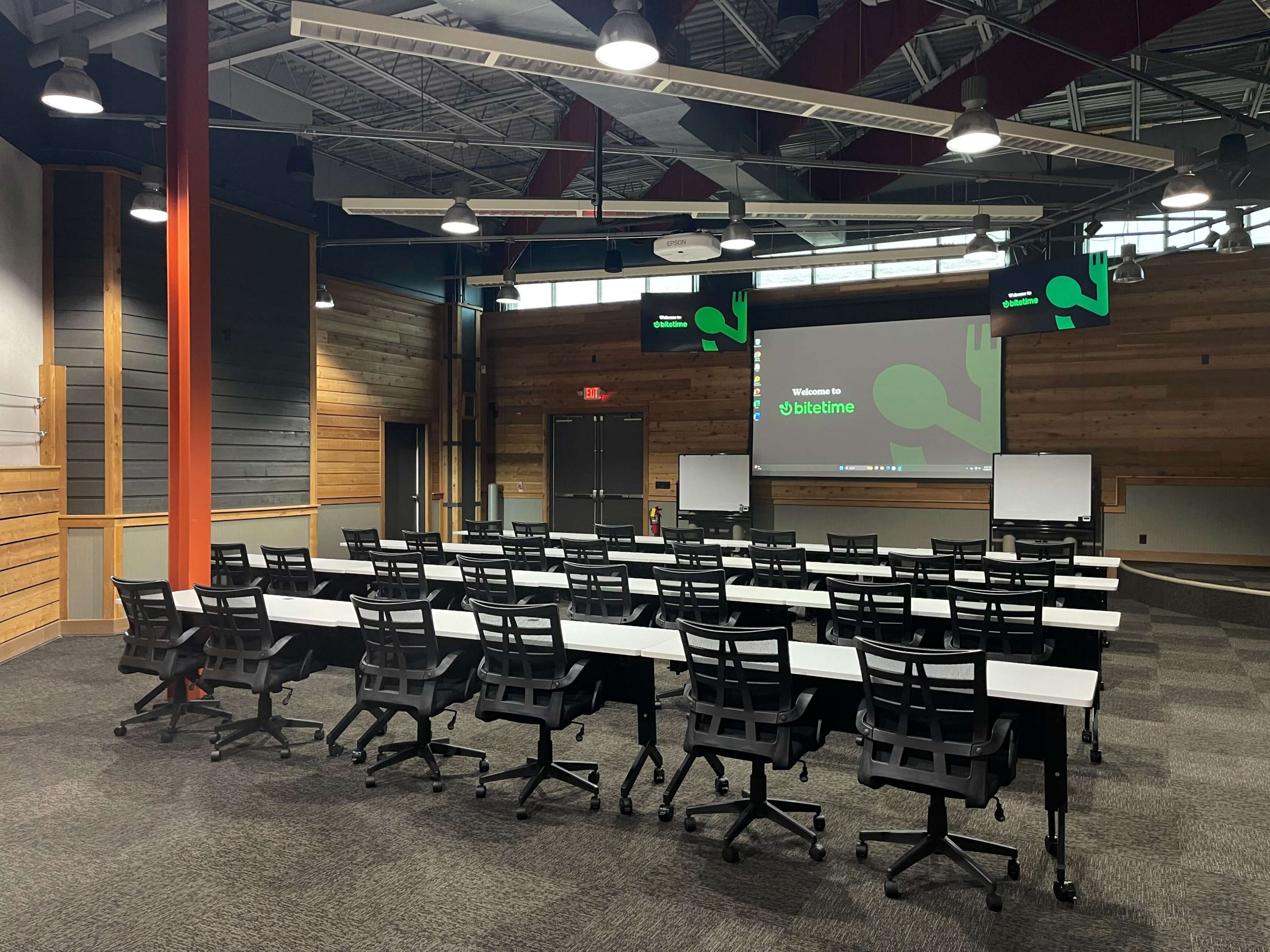 The image shows a conference room set up with rows of tables and chairs, a screen displaying a welcome message, and a modern design featuring wooden accents.