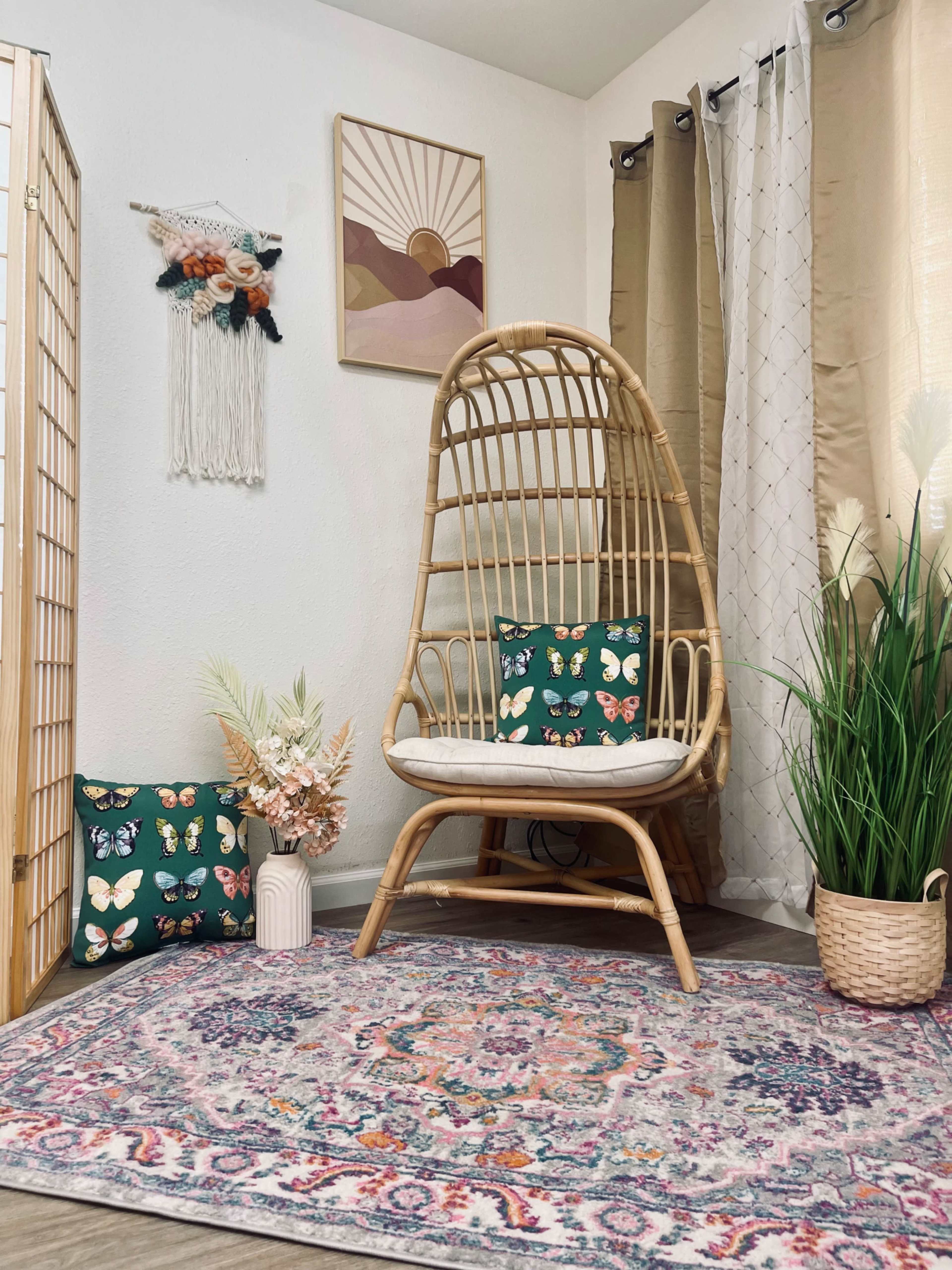 The image features a cozy corner with a rattan chair, decorative pillows, a patterned rug, and plants, set against a wall adorned with artwork and curtains.