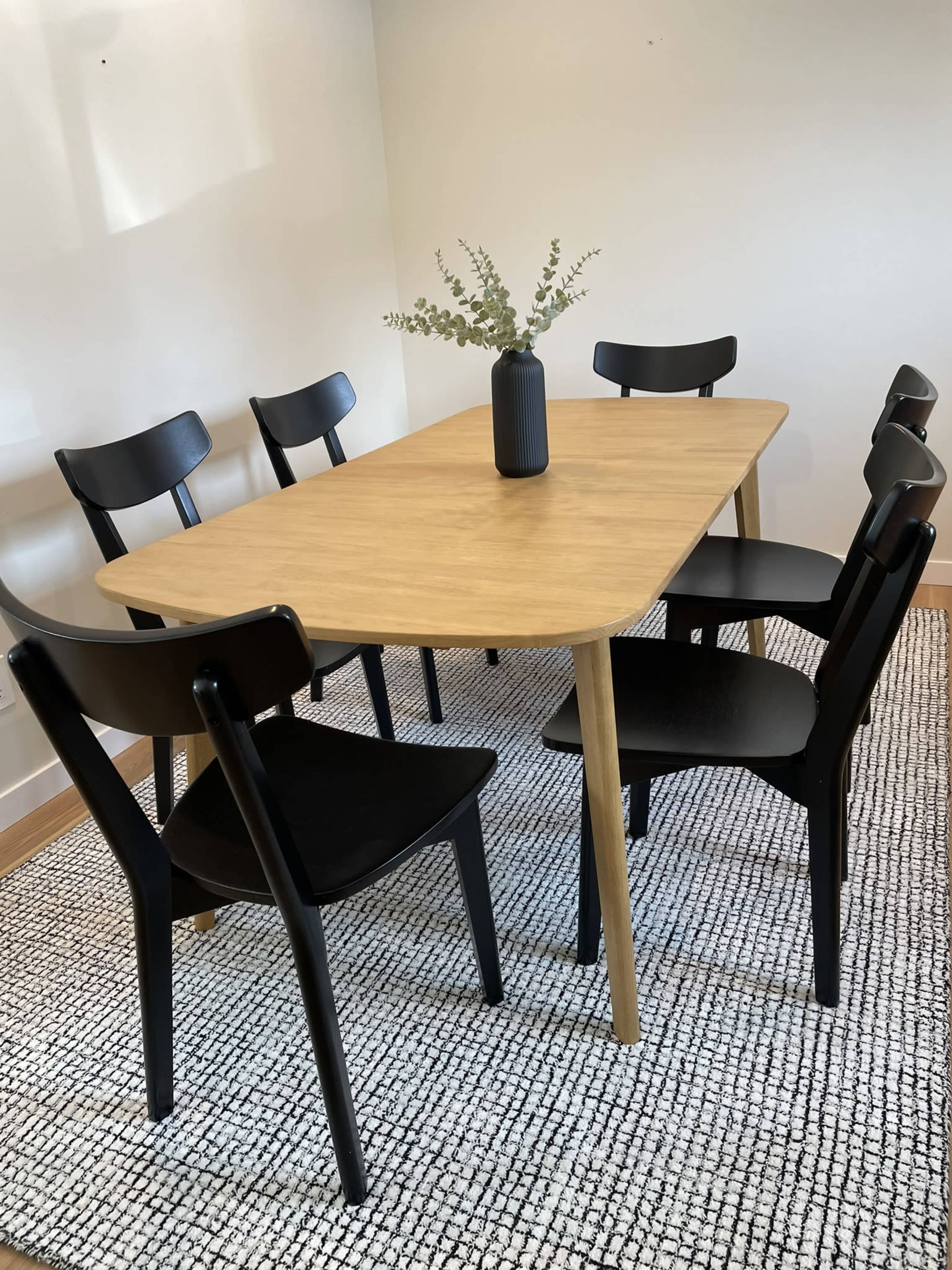 A wooden dining table surrounded by six black chairs on a textured rug with a small vase of greenery in the center.