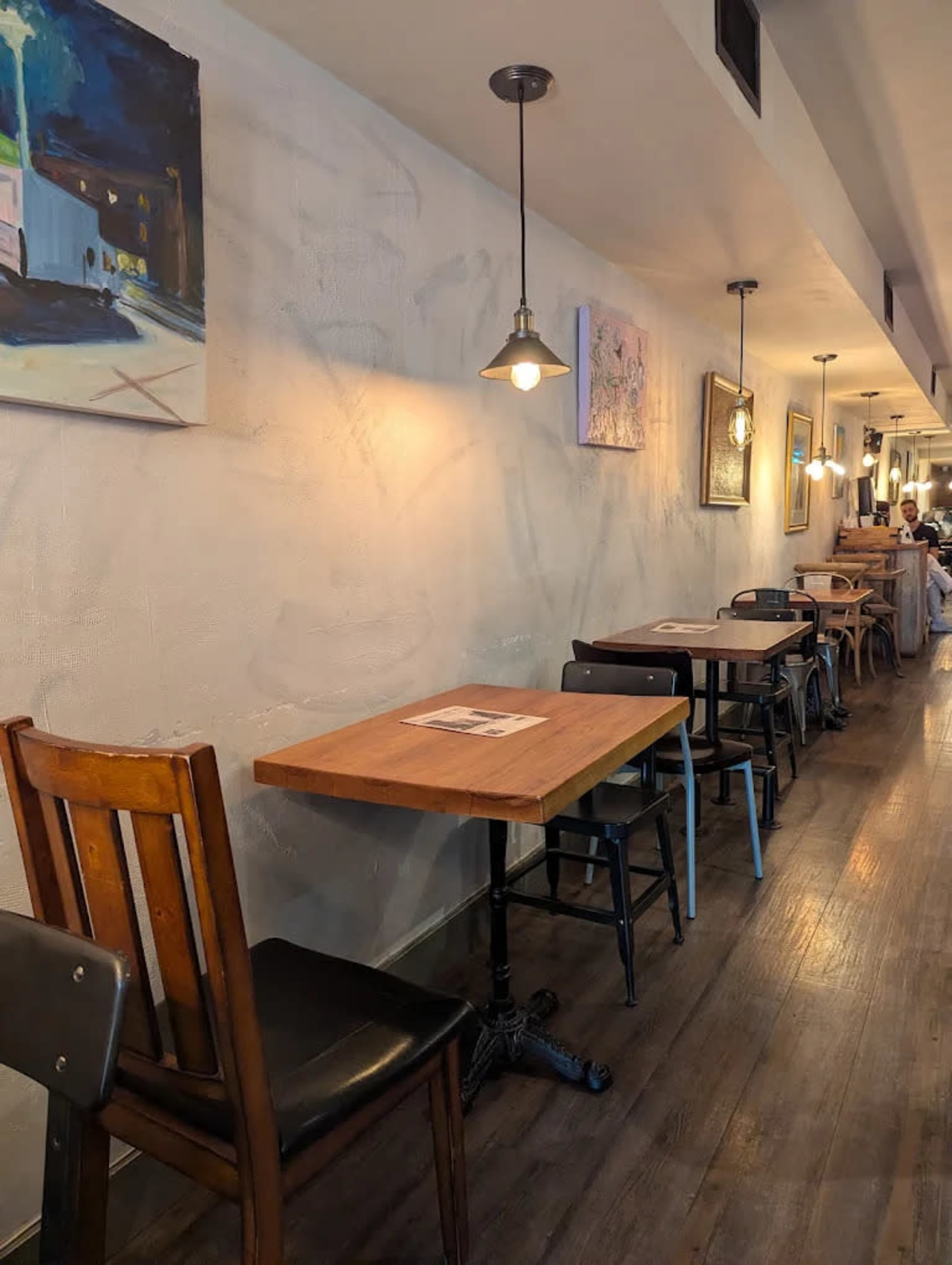 A narrow restaurant interior features a row of wooden tables and chairs along a light-colored wall, illuminated by hanging pendant lights.