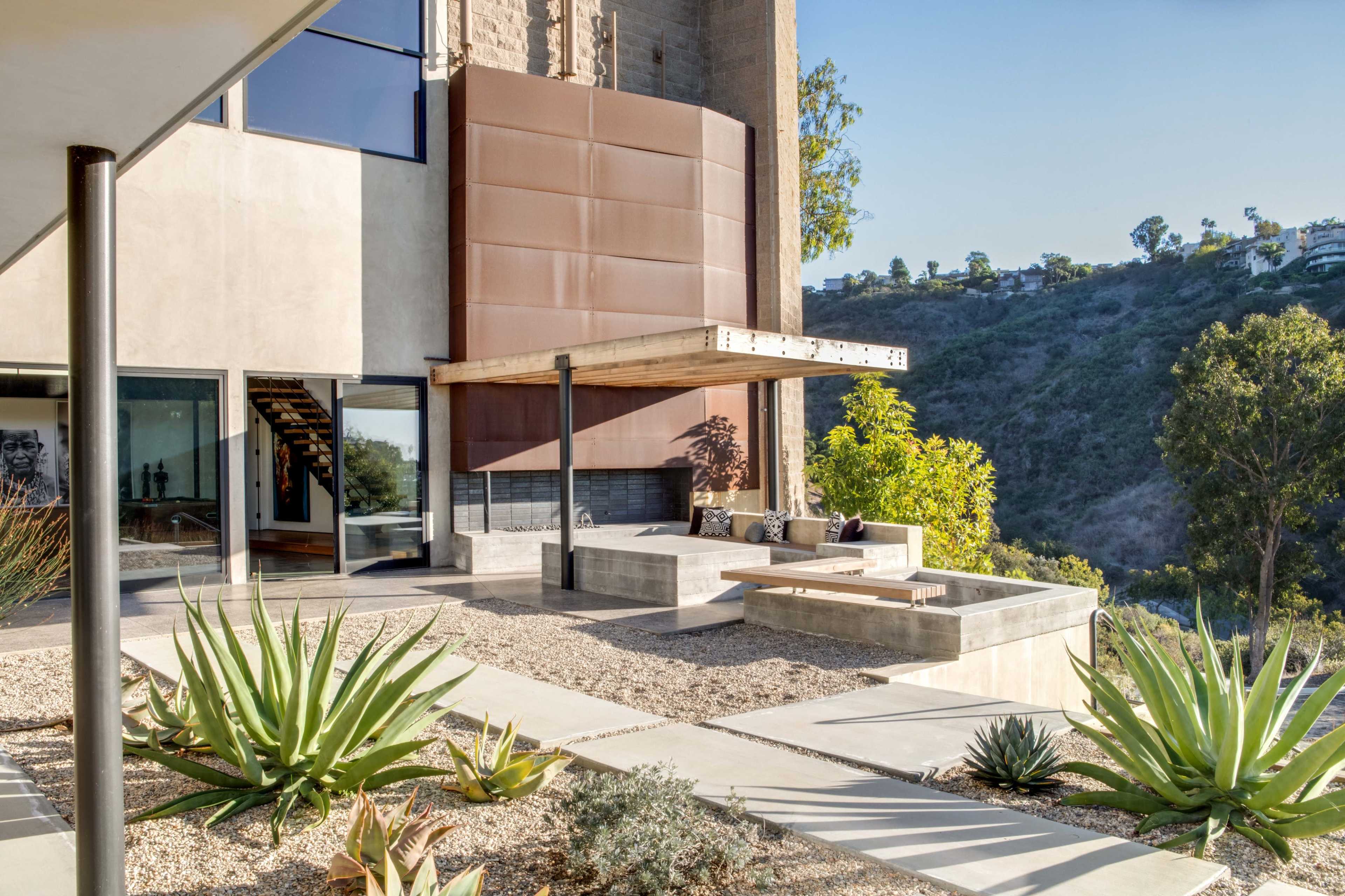 The image shows a modern patio area with a wooden overhang, seating, and landscaped outdoor space featuring succulents and a view of a hillside.