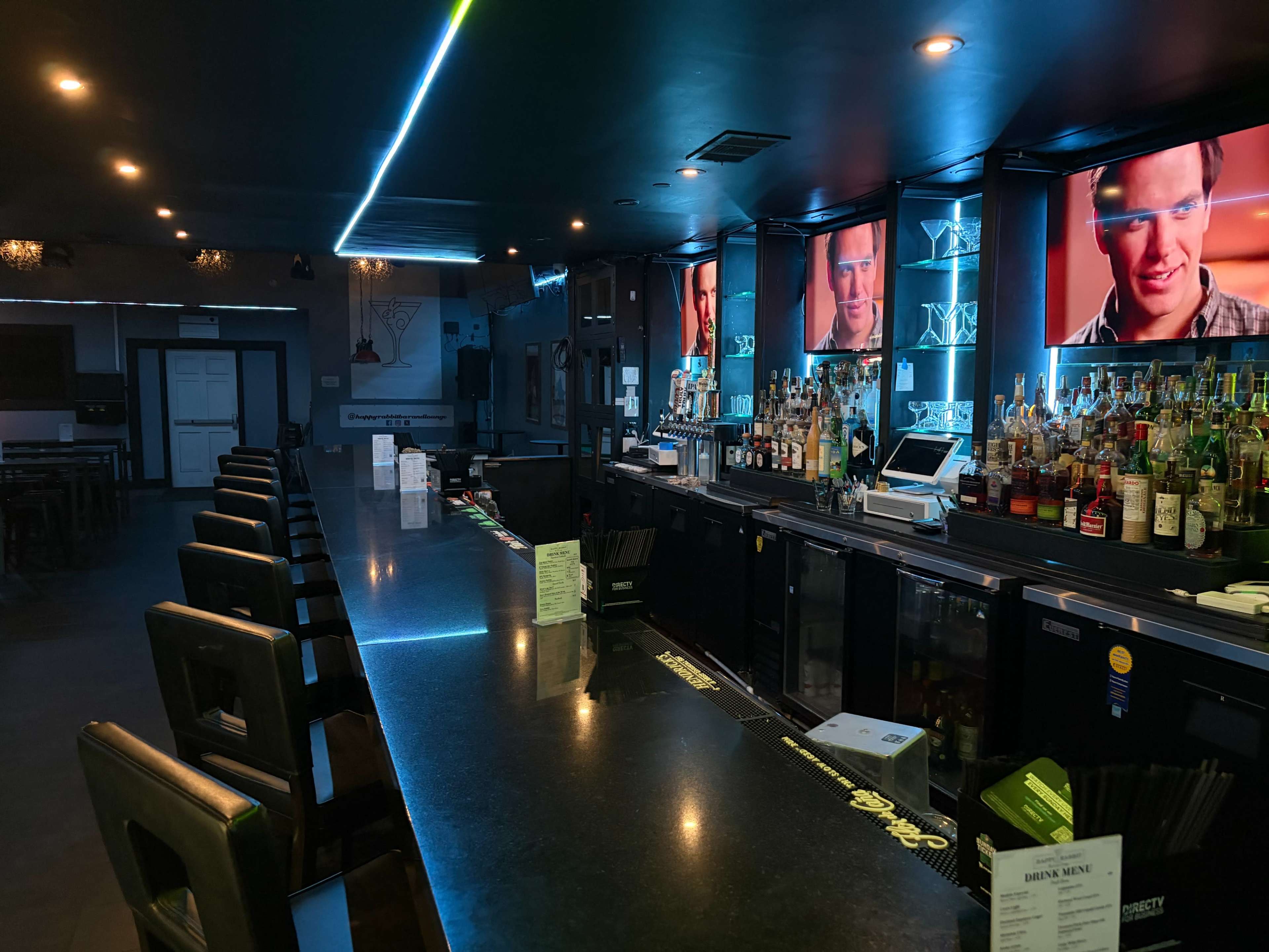 The image shows a modern bar with a long counter, illuminated by blue lighting and featuring multiple screens displaying a scene from a film.