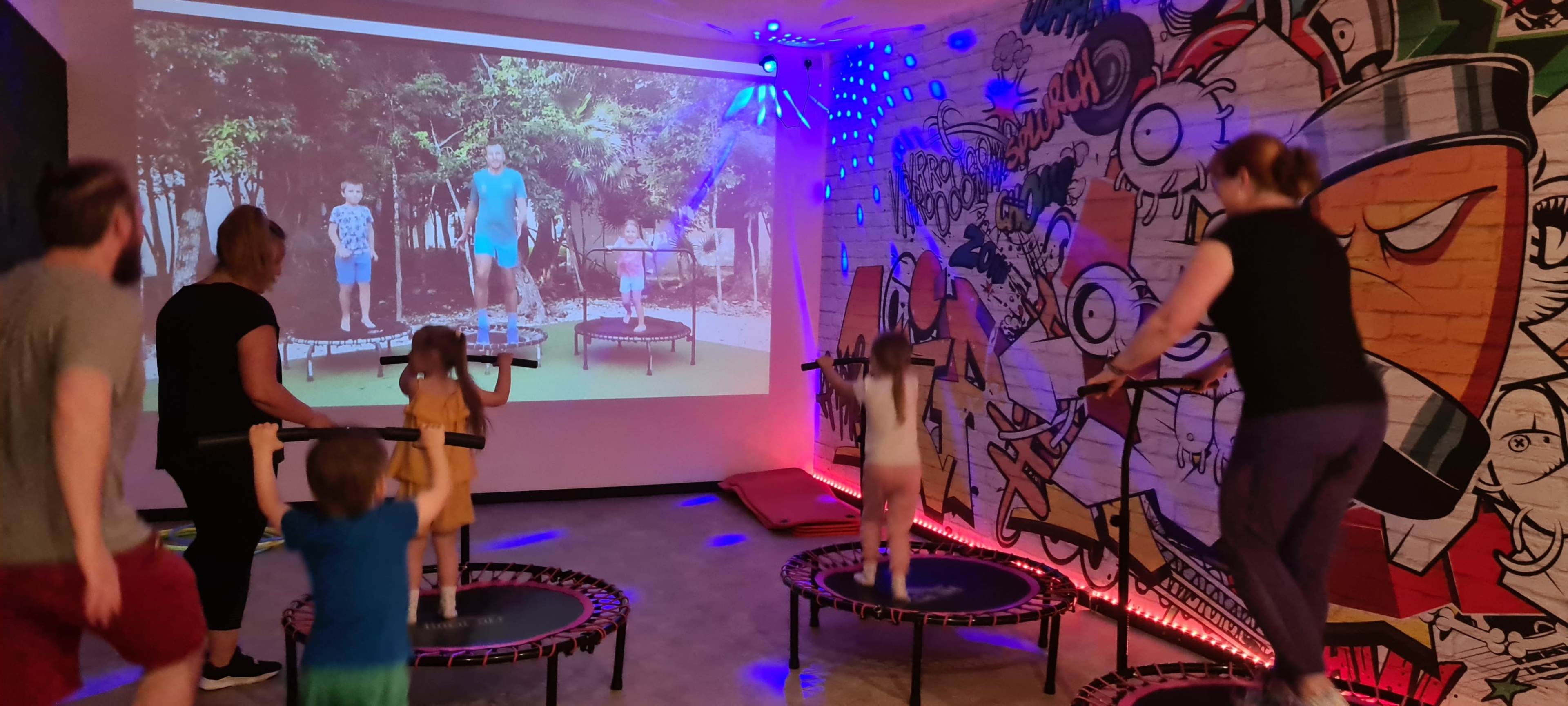 A group of adults and children jump on trampolines in a brightly lit room while a video of similar activities is projected on a wall.