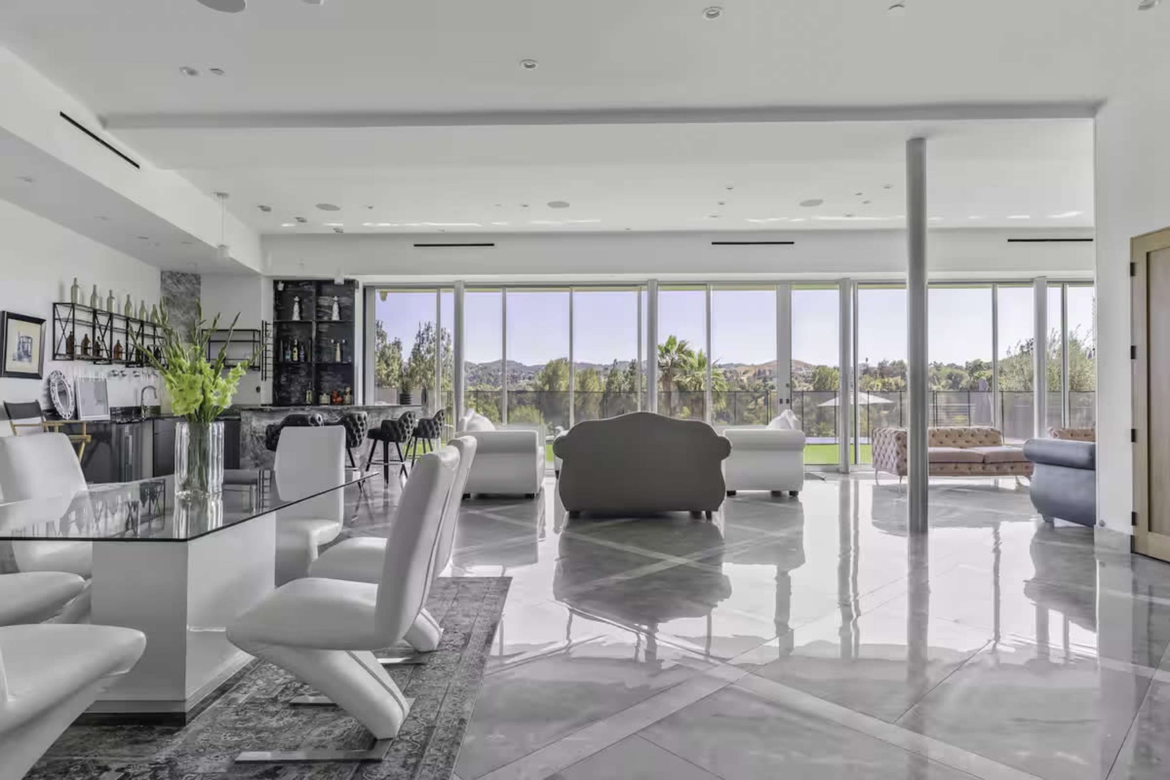 The image shows a modern, open-concept living space featuring a kitchen area with a bar, a dining table, and large windows that overlook a landscaped exterior.