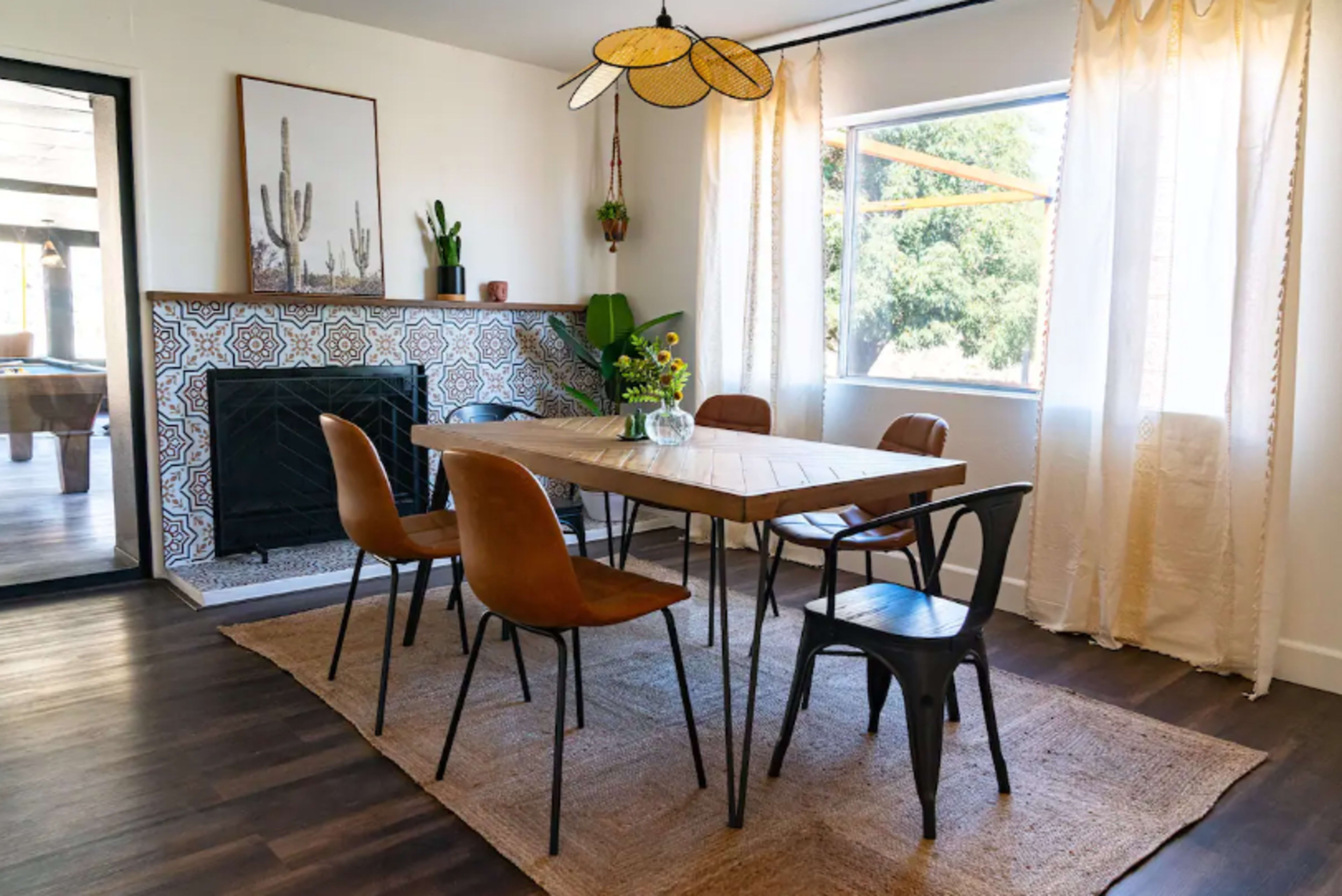 A dining area features a wooden table surrounded by six chairs, a patterned tile fireplace, and large windows with sheer curtains.