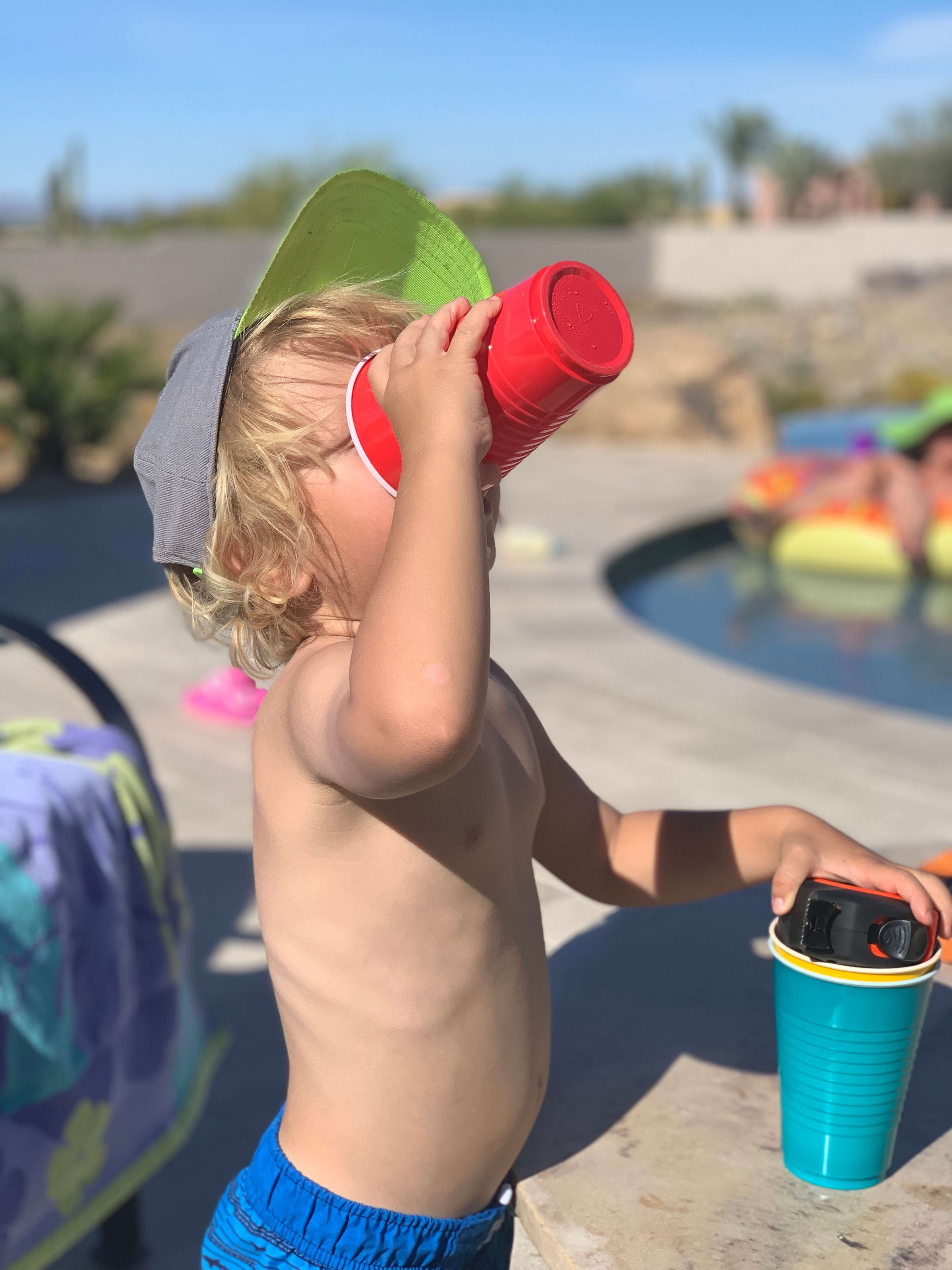 A young child wearing a cap drinks from a red cup while standing near a pool.