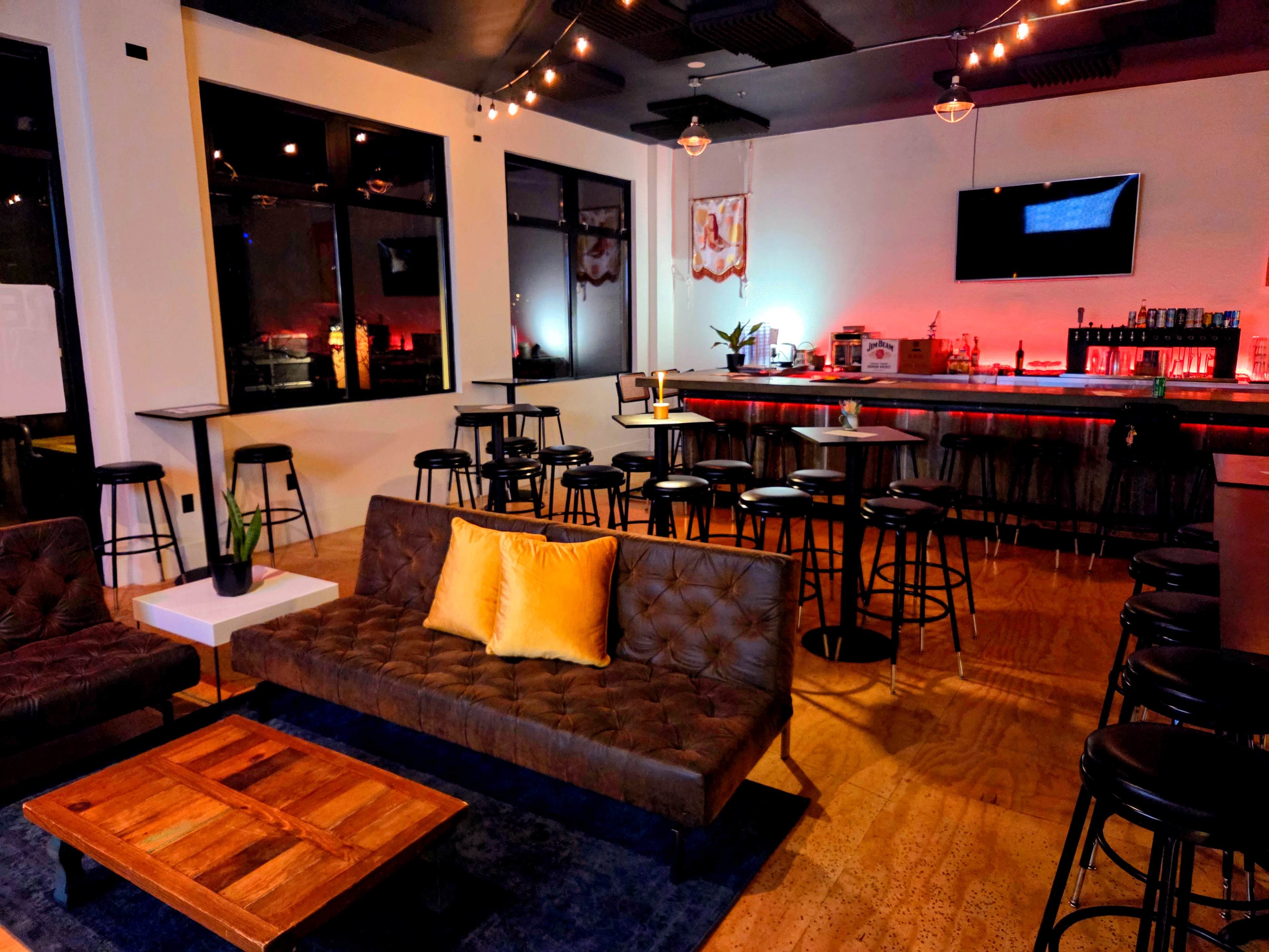 The image shows a modern lounge area with a dark leather sofa, wooden coffee table, and bar space illuminated by dim lighting and decorative accents.