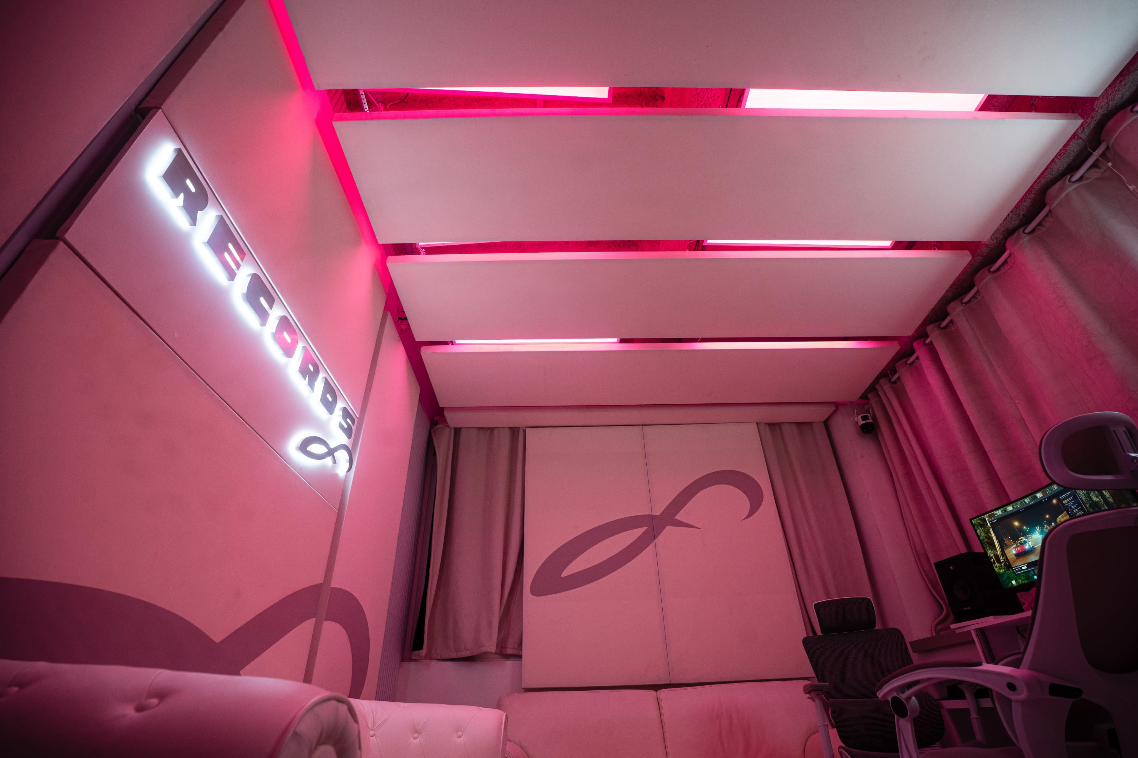 The image shows a modern recording studio with pink lighting, white walls, and a computer setup.
