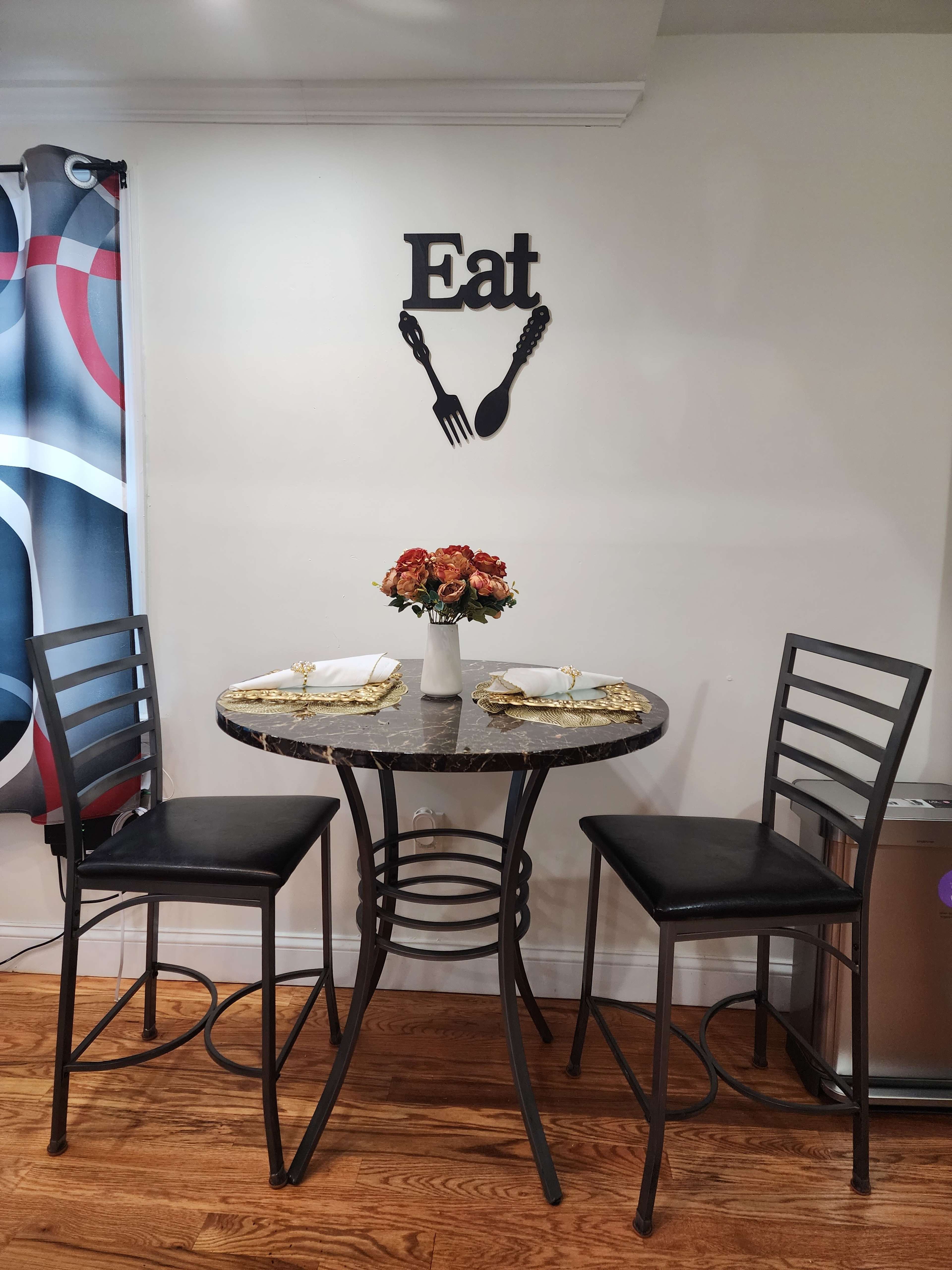 A small dining table with two black chairs is set against a wall adorned with a decorative "Eat" sign and a vase of flowers on the table.