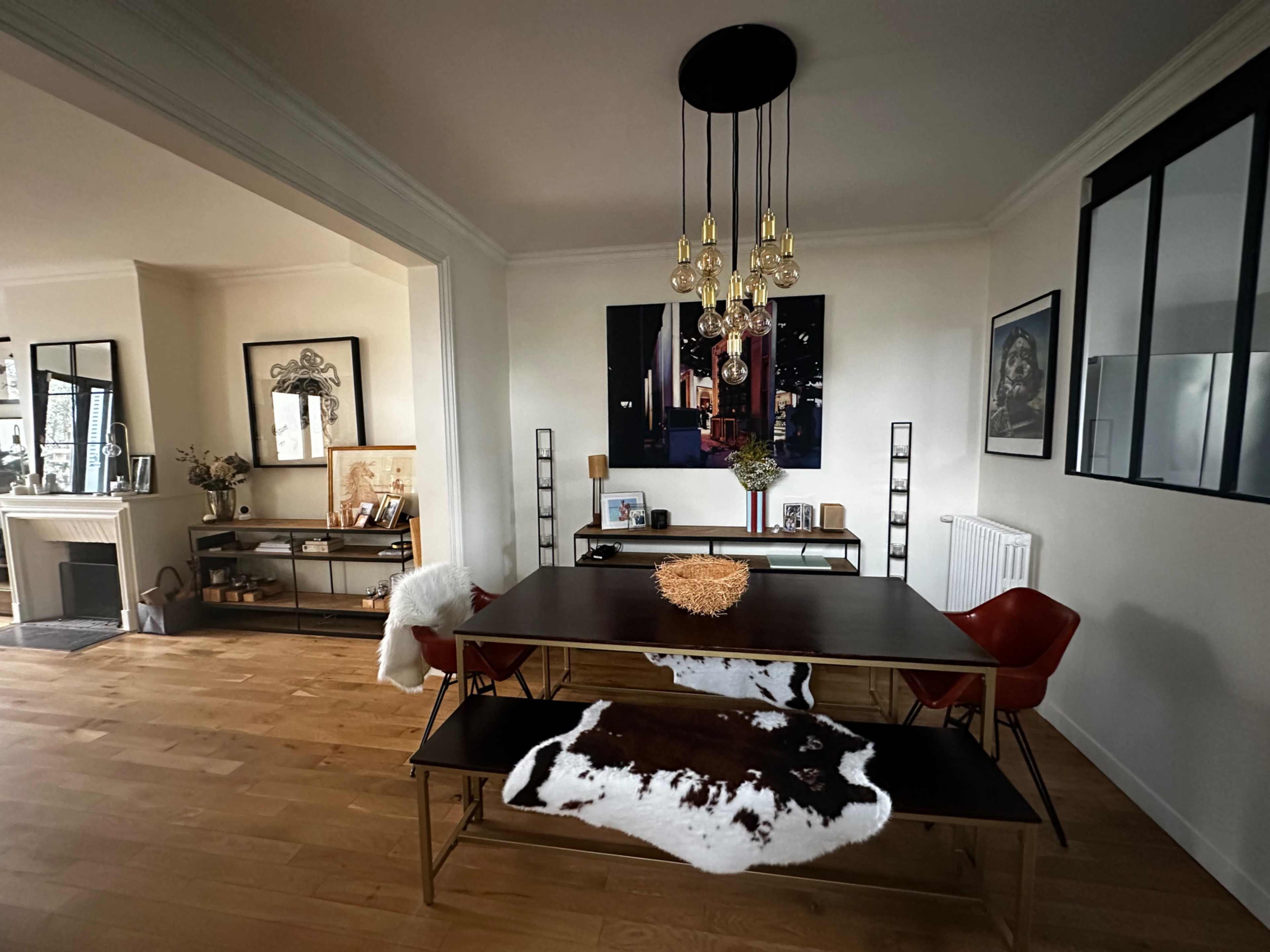 A dining area features a large wooden table with metal legs, accent chairs, and a cowhide rug, illuminated by a modern chandelier.