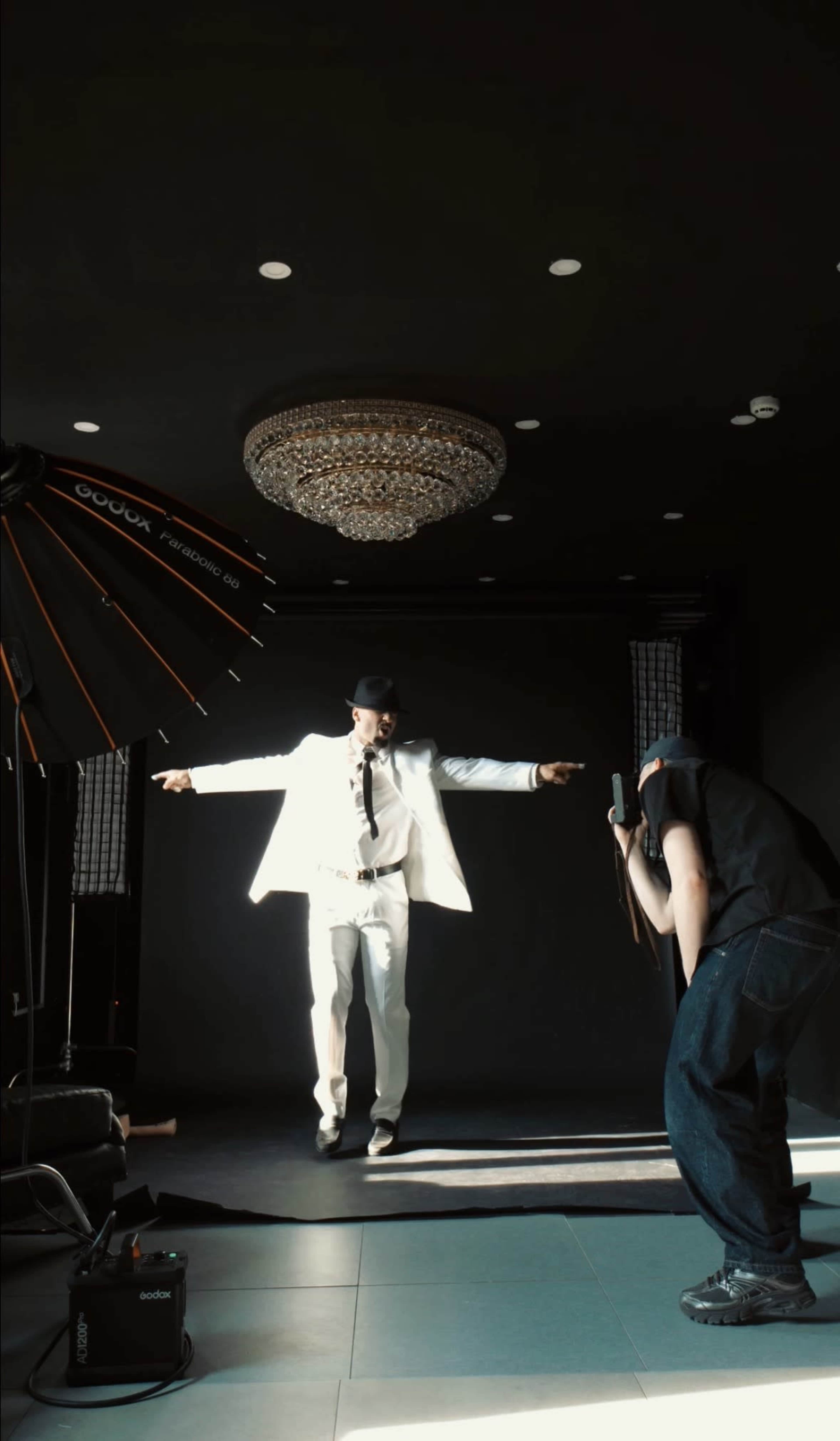 A man in a white suit and fedora poses dramatically in a photo studio while a photographer captures the shot.