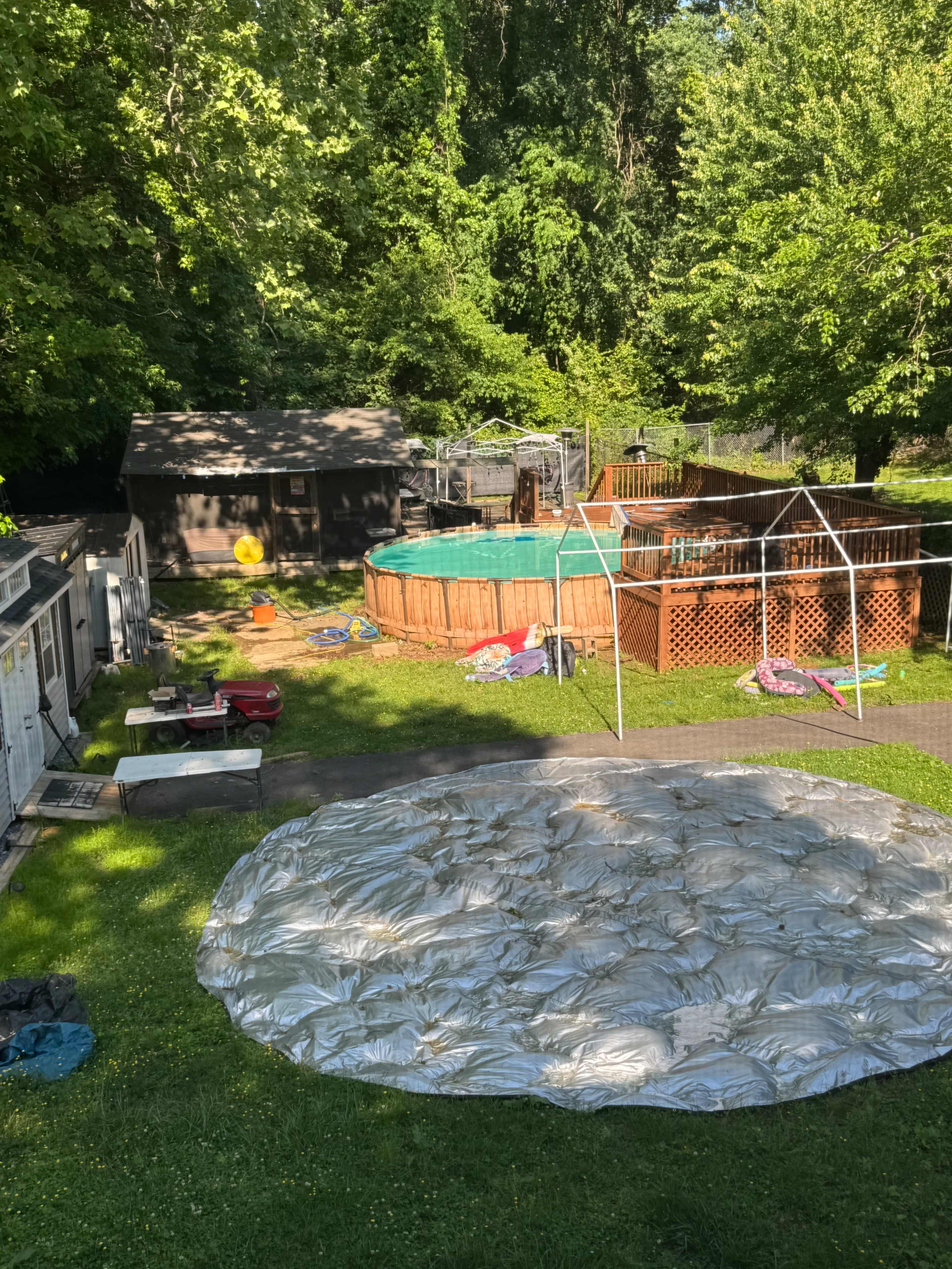 The image shows a backyard with a swimming pool, a large silver tarp laid out on the grass, picnic tables, and several inflatable toys scattered around.