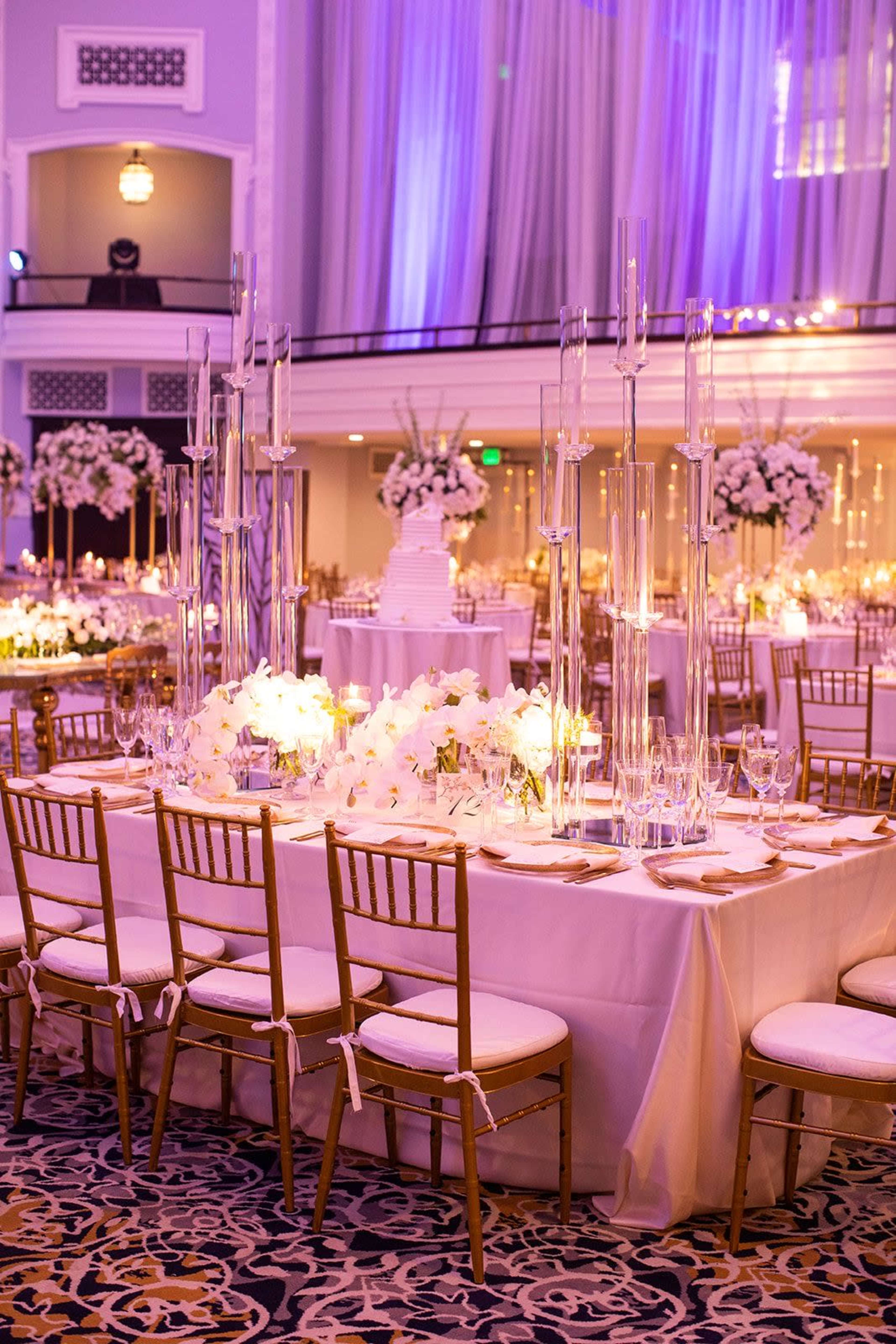 The image shows an elegantly set banquet hall with long tables adorned with floral arrangements and tall candle holders, all under soft purple lighting.