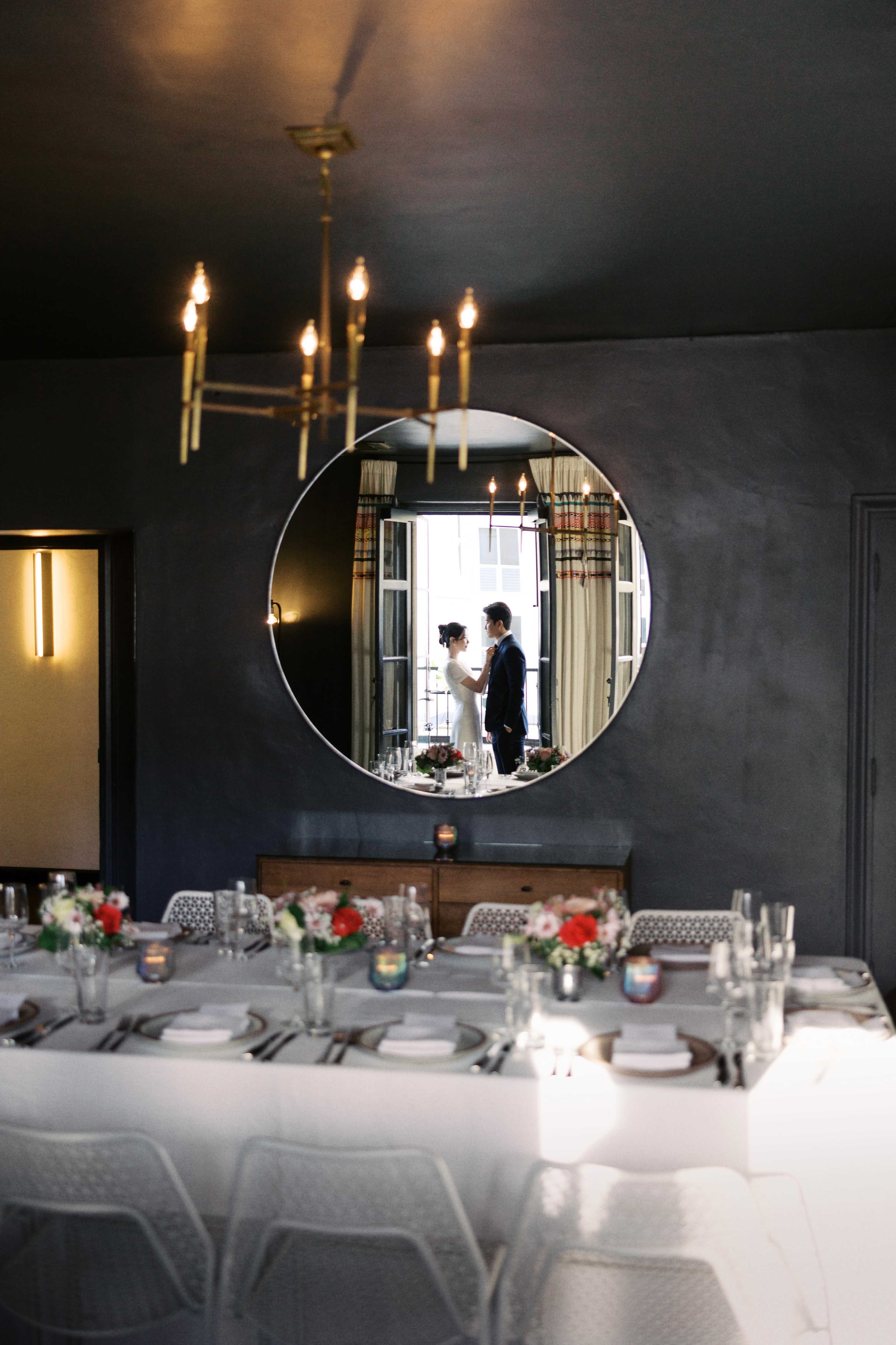 A couple stands together in a dimly lit room reflected in a round mirror, with a decorated dining table set for a celebration in the foreground.