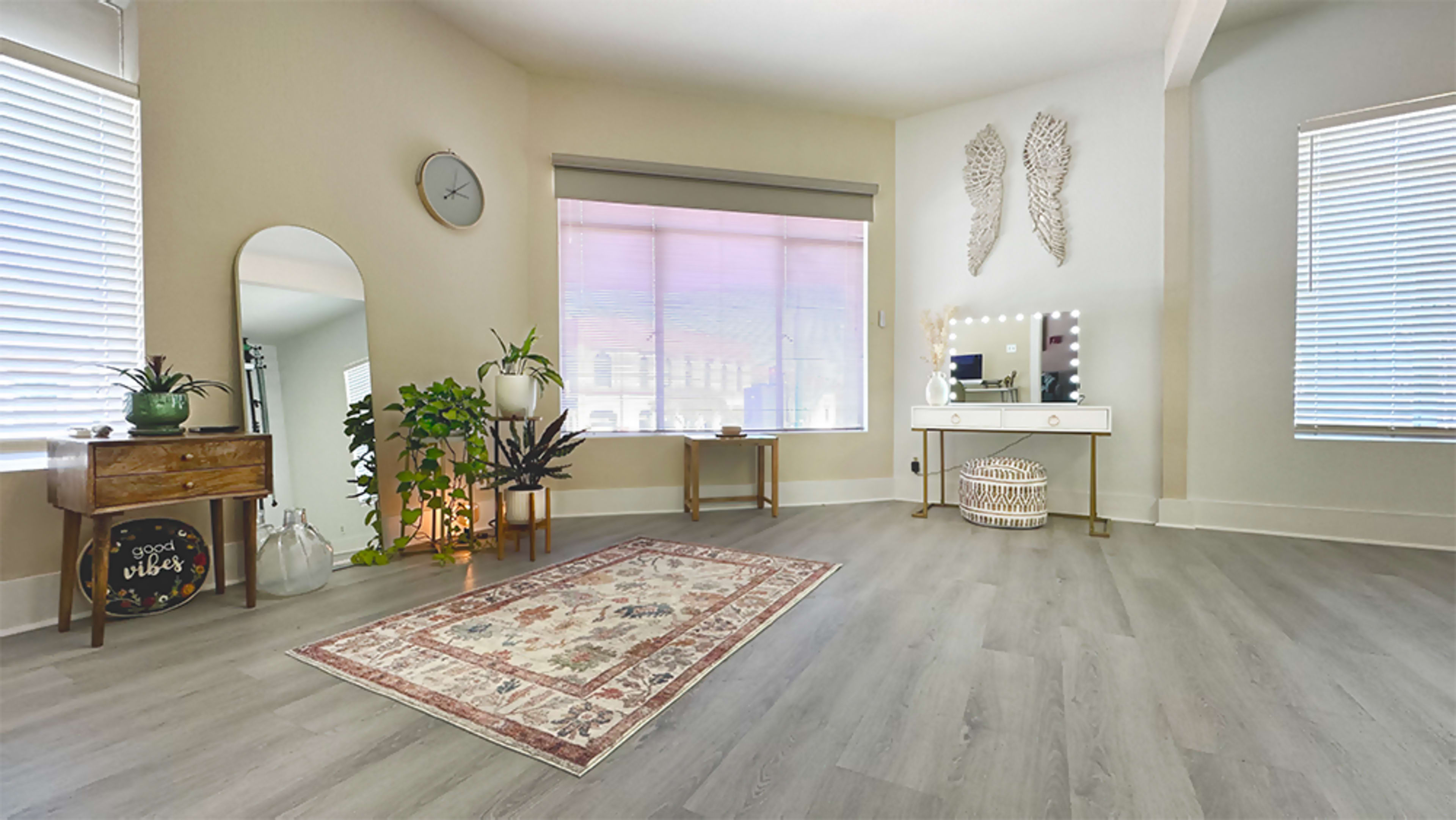 A spacious, well-lit room with a decorative rug, a vanity table, a large mirror, plants, and a window with blinds.