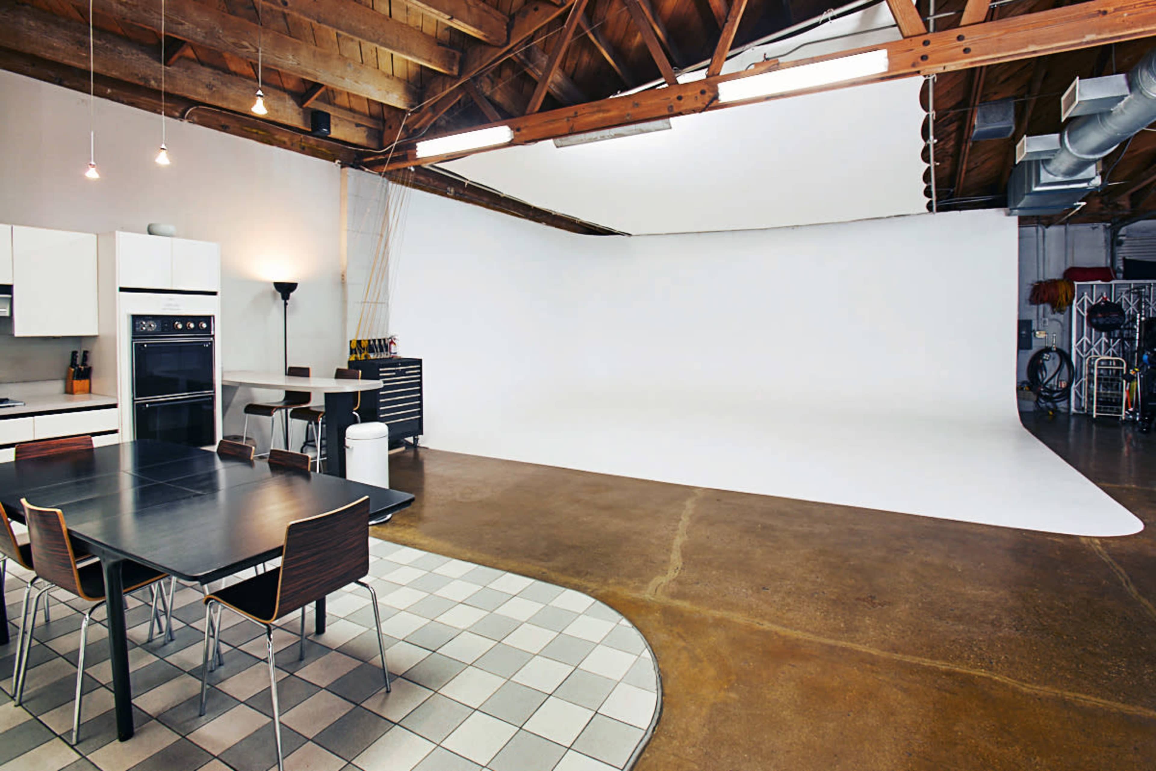 A modern kitchen and dining area adjoins a spacious room with a large white backdrop, featuring hardwood beams and a concrete floor.