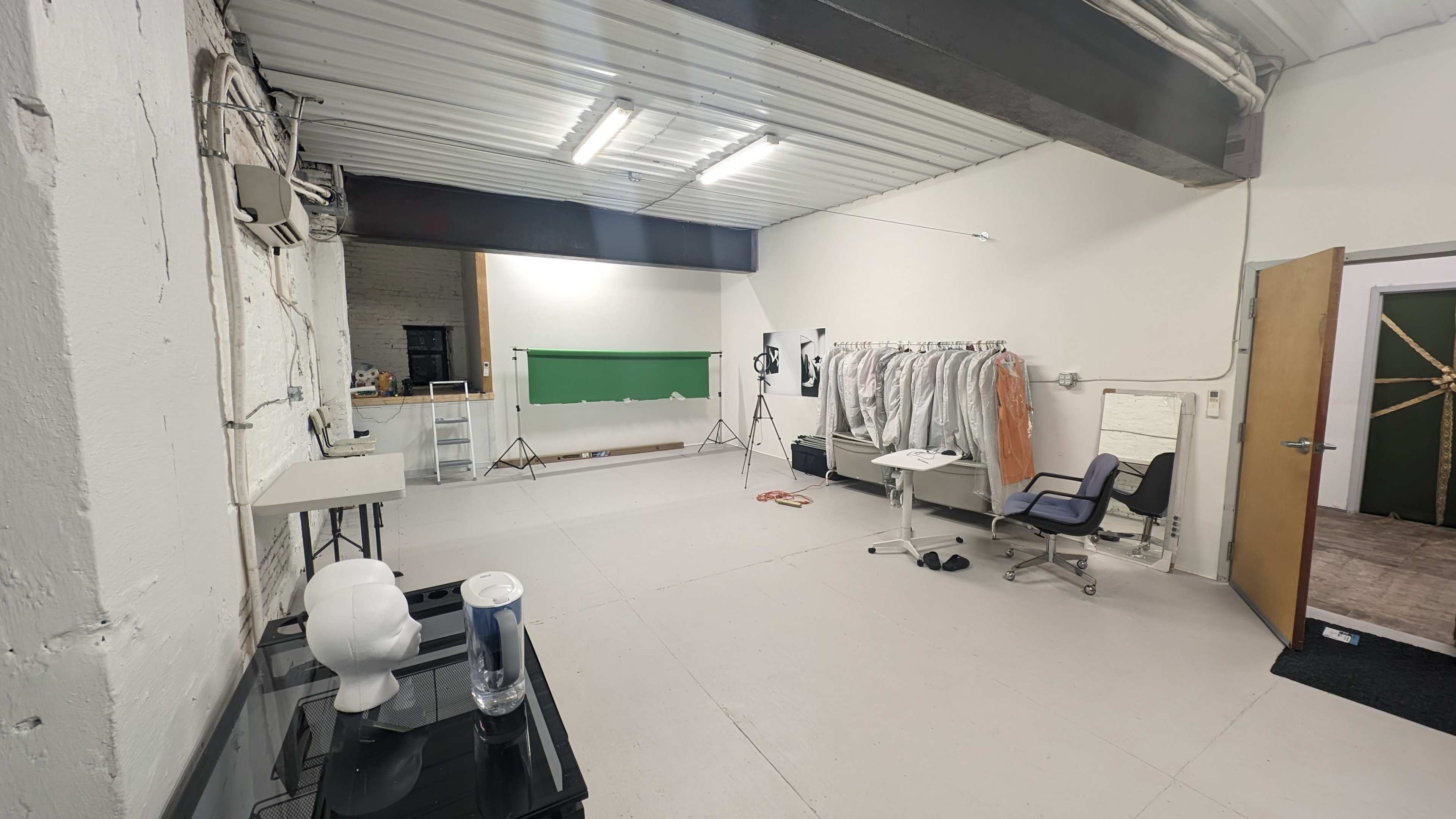 The image shows a spacious studio with a green backdrop, a clothing rack filled with garments, a desk with a chair, and a mannequin on a table.