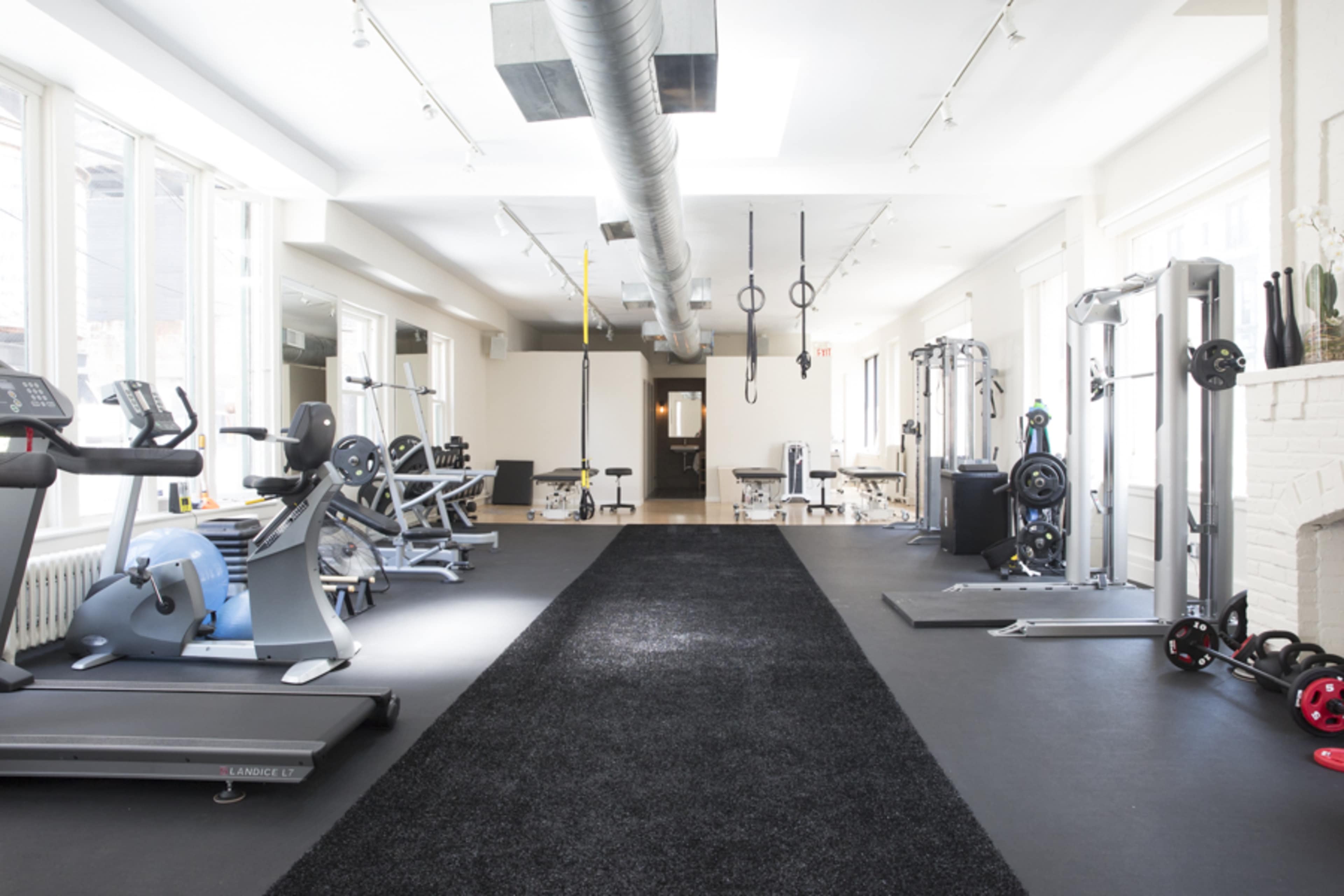 The image shows a spacious gym featuring various workout machines, cardio equipment, and free weights arranged along a central black carpeted pathway.