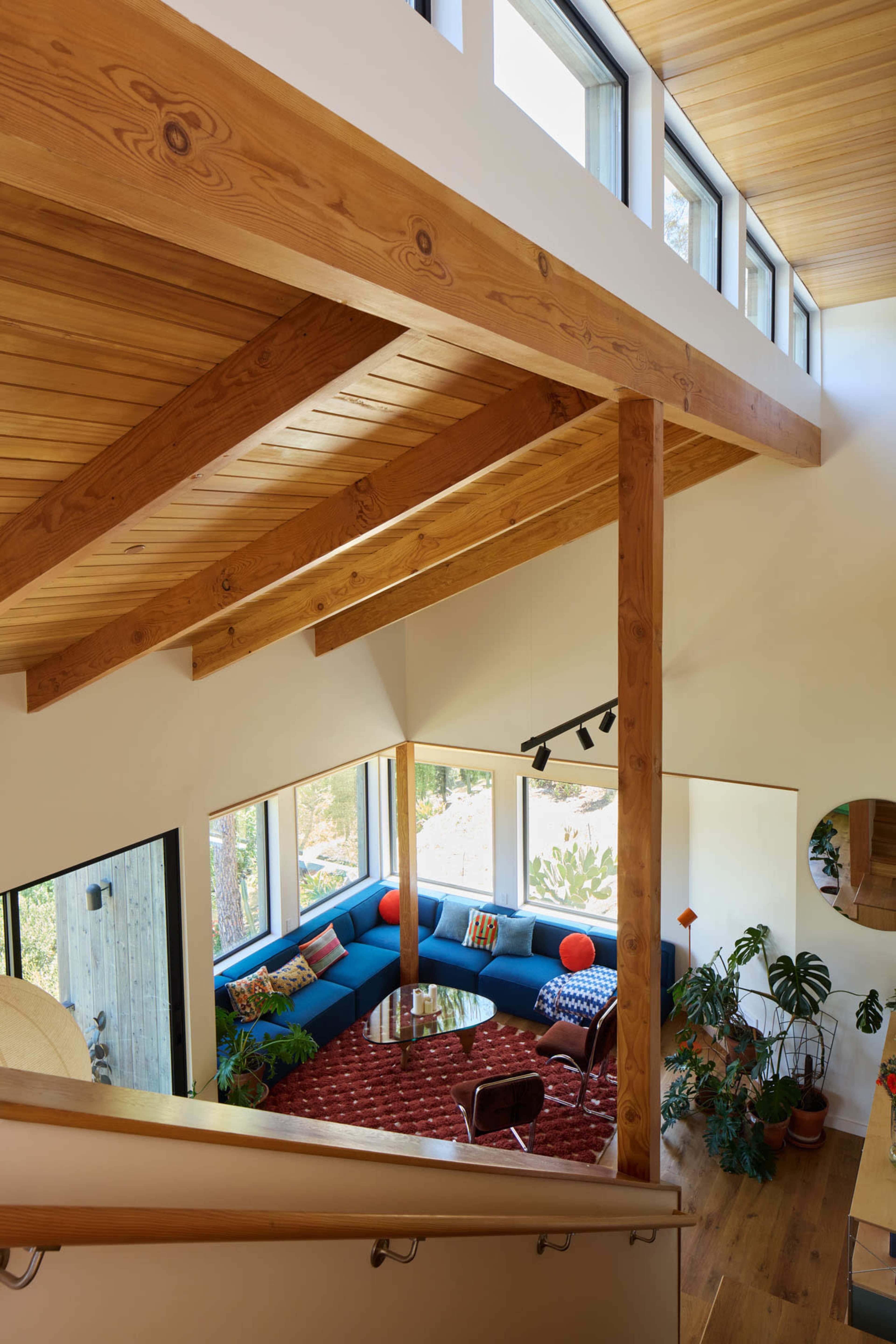 The image shows a modern living space with wooden beams, a blue sectional sofa, and large windows allowing natural light to flood the room, adorned with potted plants and decorative cushions.