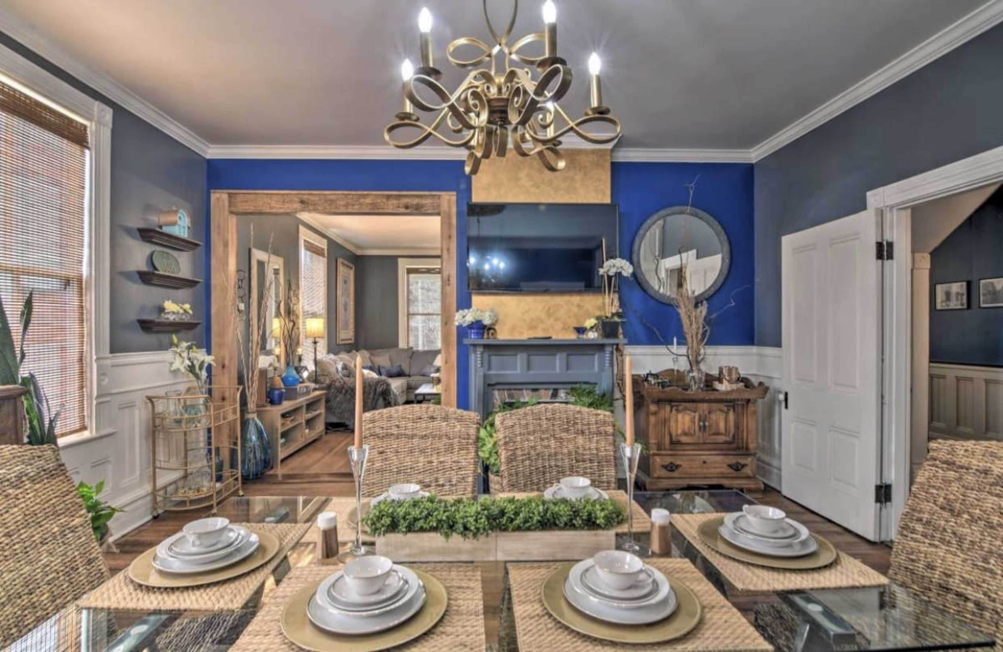 A dining room features a glass table set with plates and greenery, leading into a living area with a blue accent wall and a mounted television.