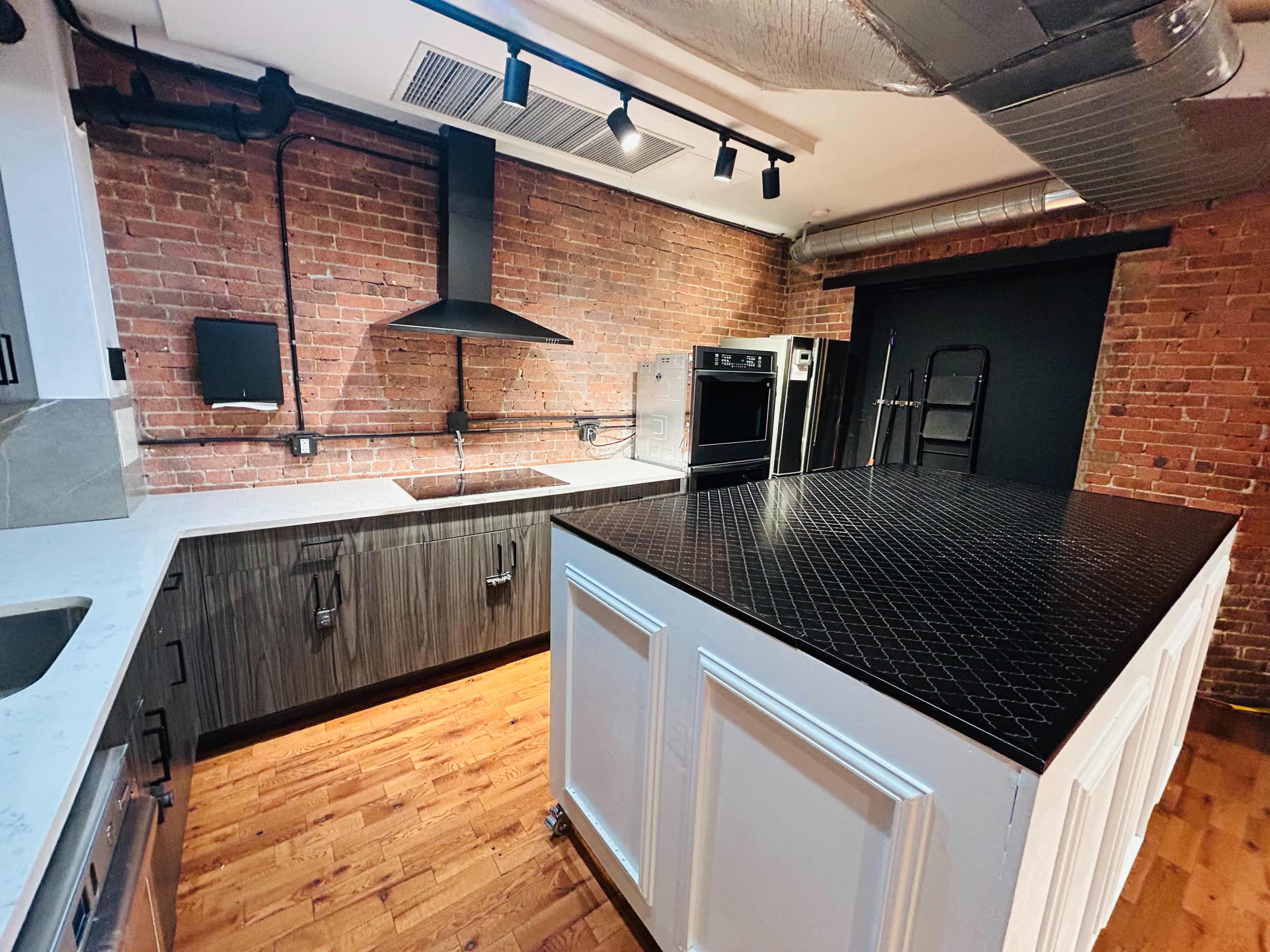 The image shows a modern kitchen featuring exposed brick walls, a large island with a black patterned countertop, and stainless steel appliances.