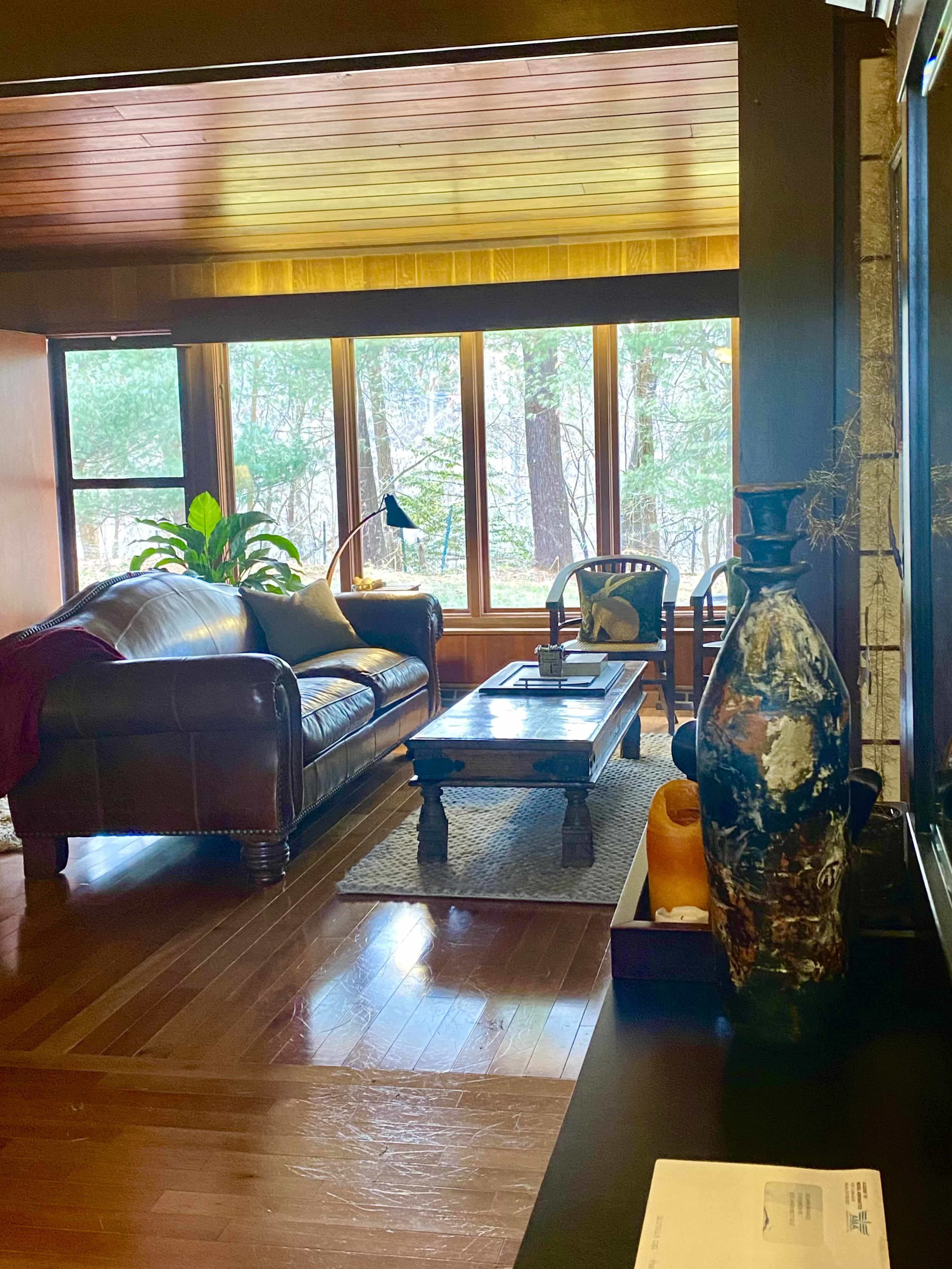 A cozy living room with a leather sofa, a wooden coffee table, and large windows that provide a view of a wooded area outside.